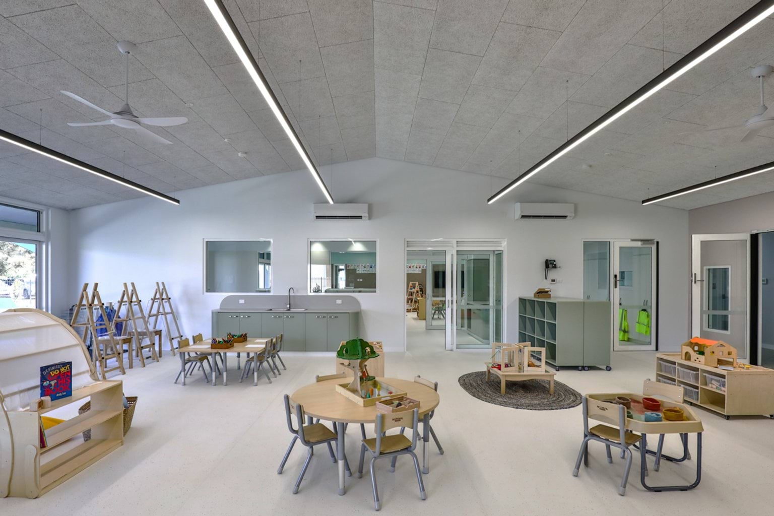 A kindergarten classroom with small round tables, timber storage units, easels, and a reading nook near the windows.