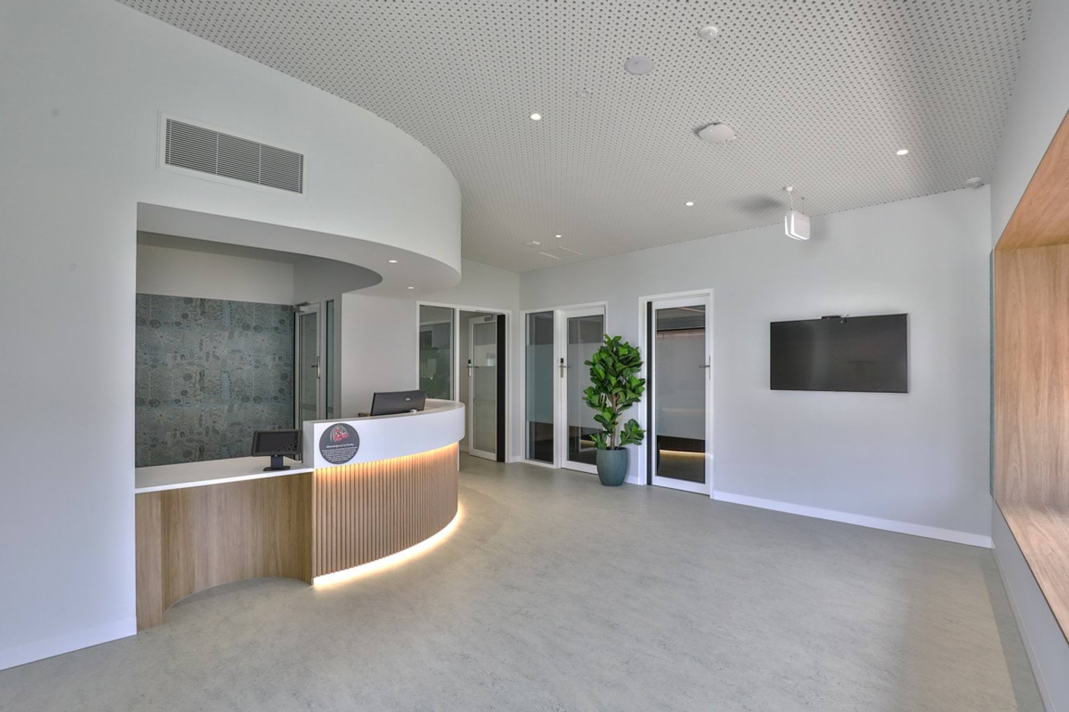 A school reception area with a curved desk, light timber finishes, and adjoining office rooms, with a large screen and indoor plant.