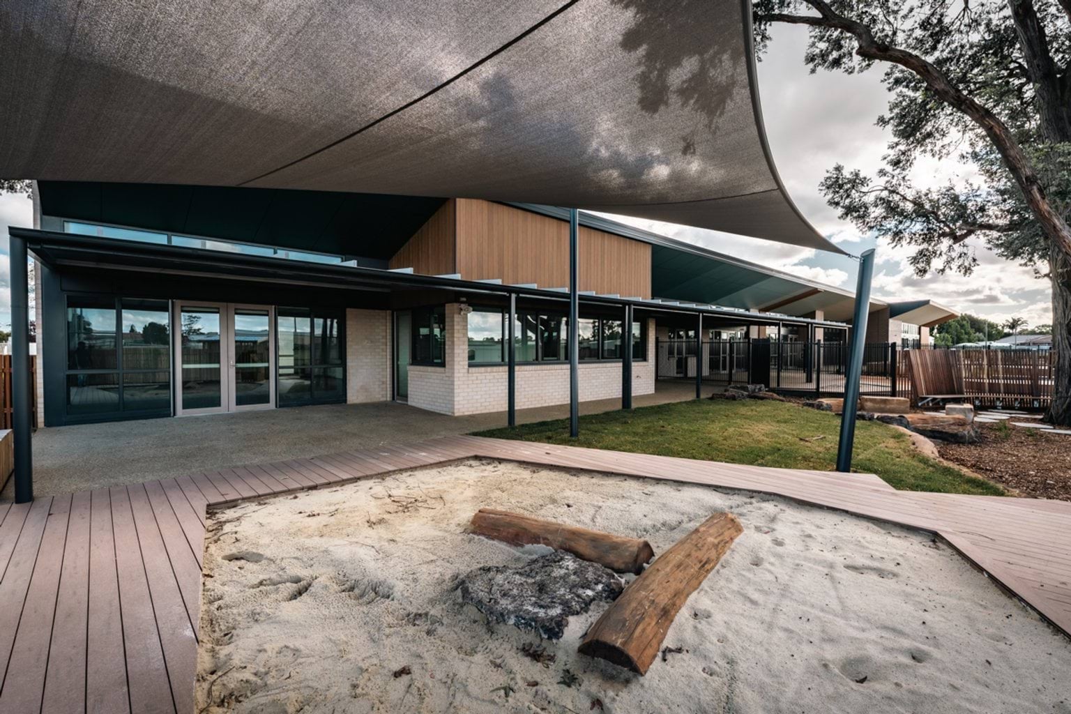 An outdoor kindergarten play space with a shaded deck, sandpit with logs, and landscaped areas beside the building.