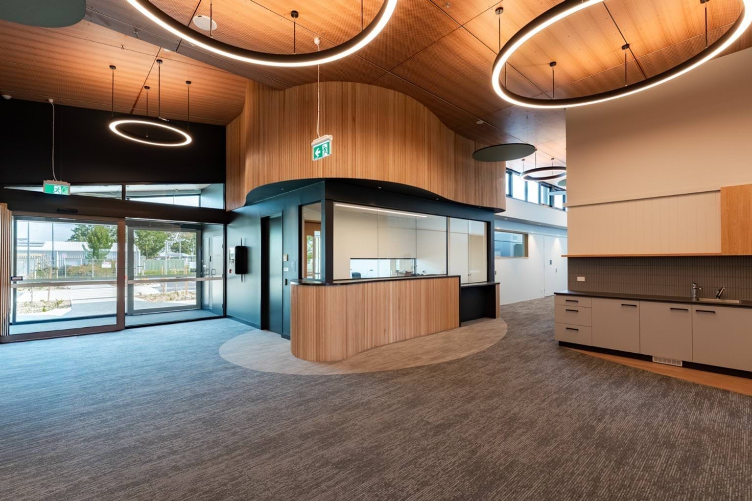 A reception space with timber ceiling curves, ring-shaped lights, and a central counter with glass partitions.
