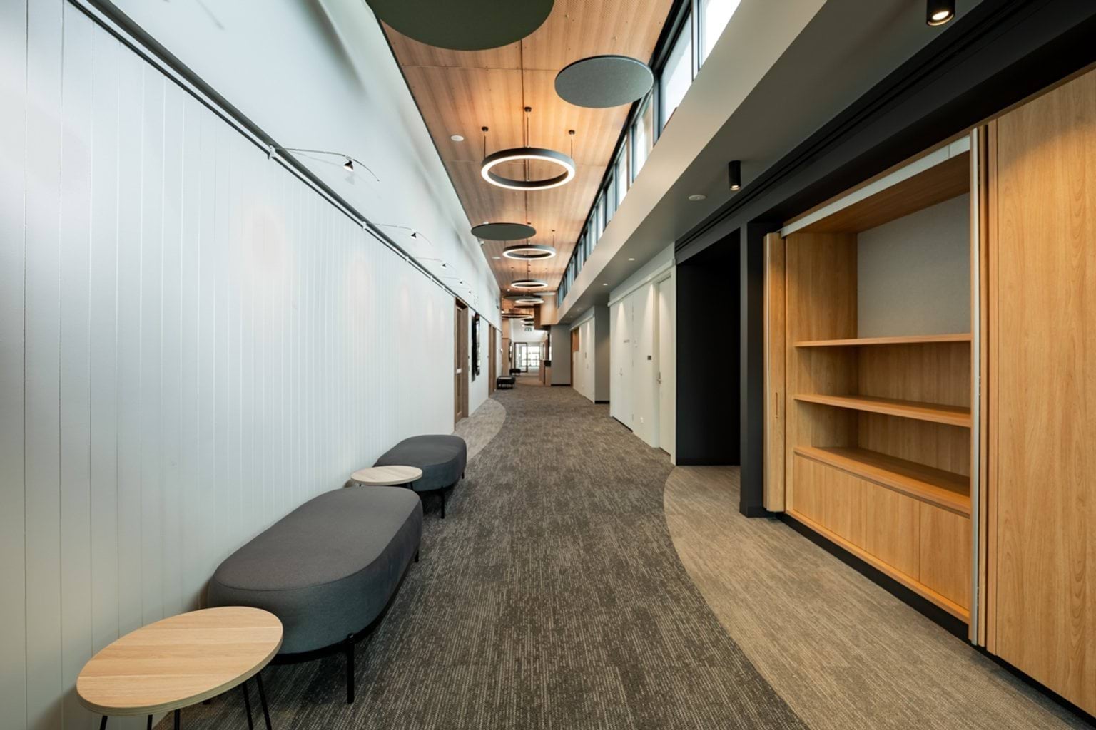 A long kindergarten corridor with cushioned seating, timber ceiling panels, circular light fittings, and open shelving.
