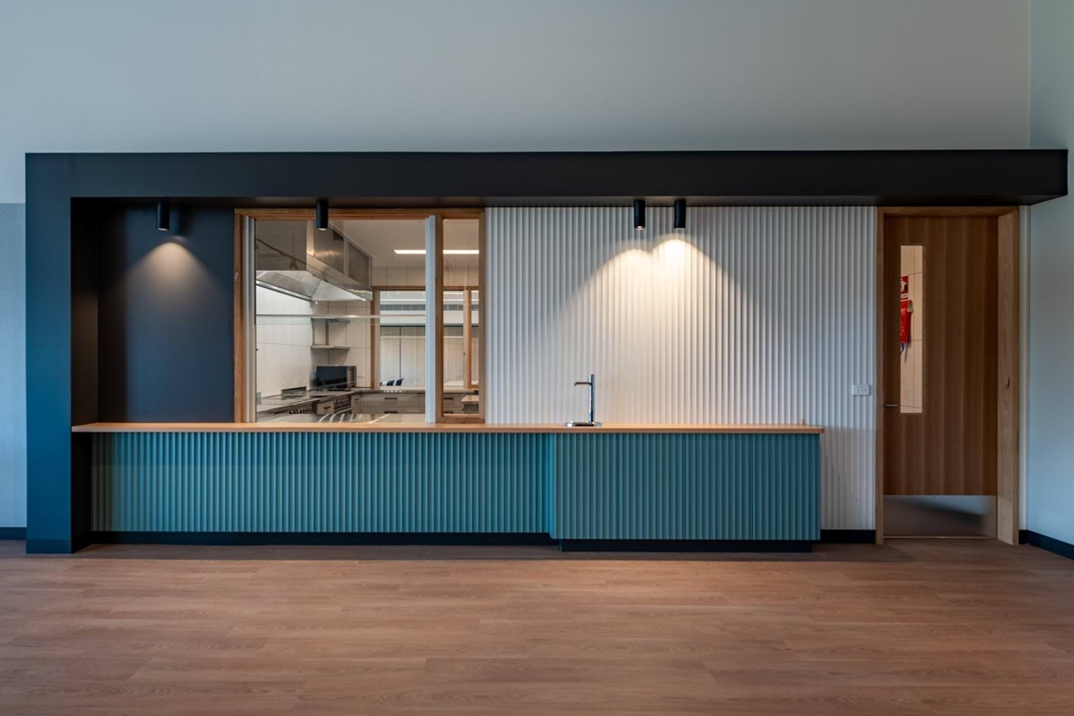 A kitchenette with teal cabinetry, ribbed panelling, a timber benchtop, and a glass viewing window into an adjacent room.