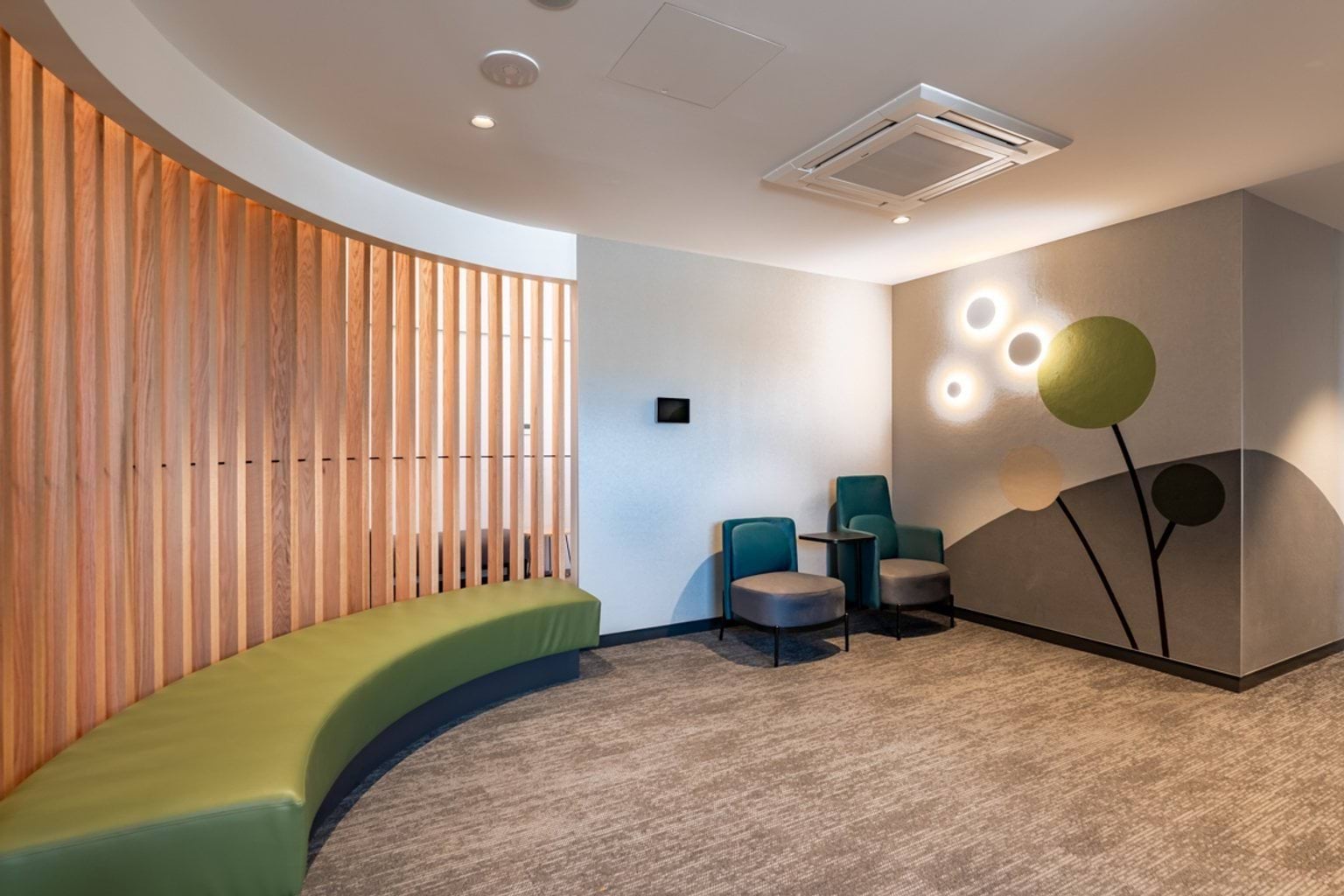 A calm waiting area with curved green seating, timber slats, and a wall mural featuring stylised trees and soft lighting.