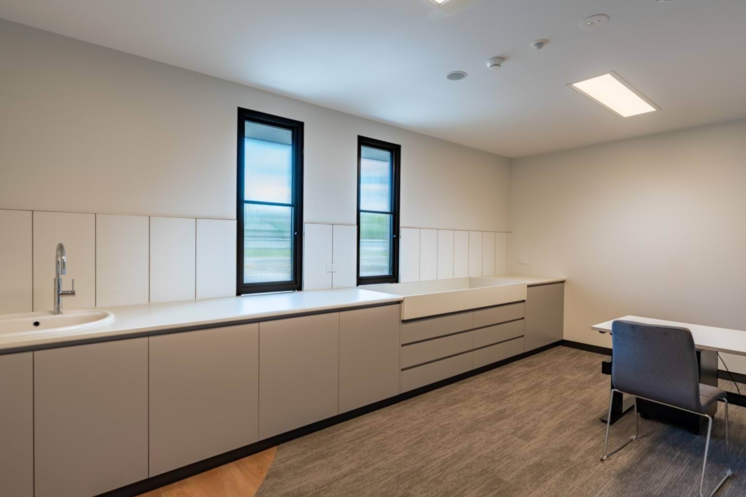 A staff or consultation room with white cabinetry, long benchtops, and two tall windows providing natural light.