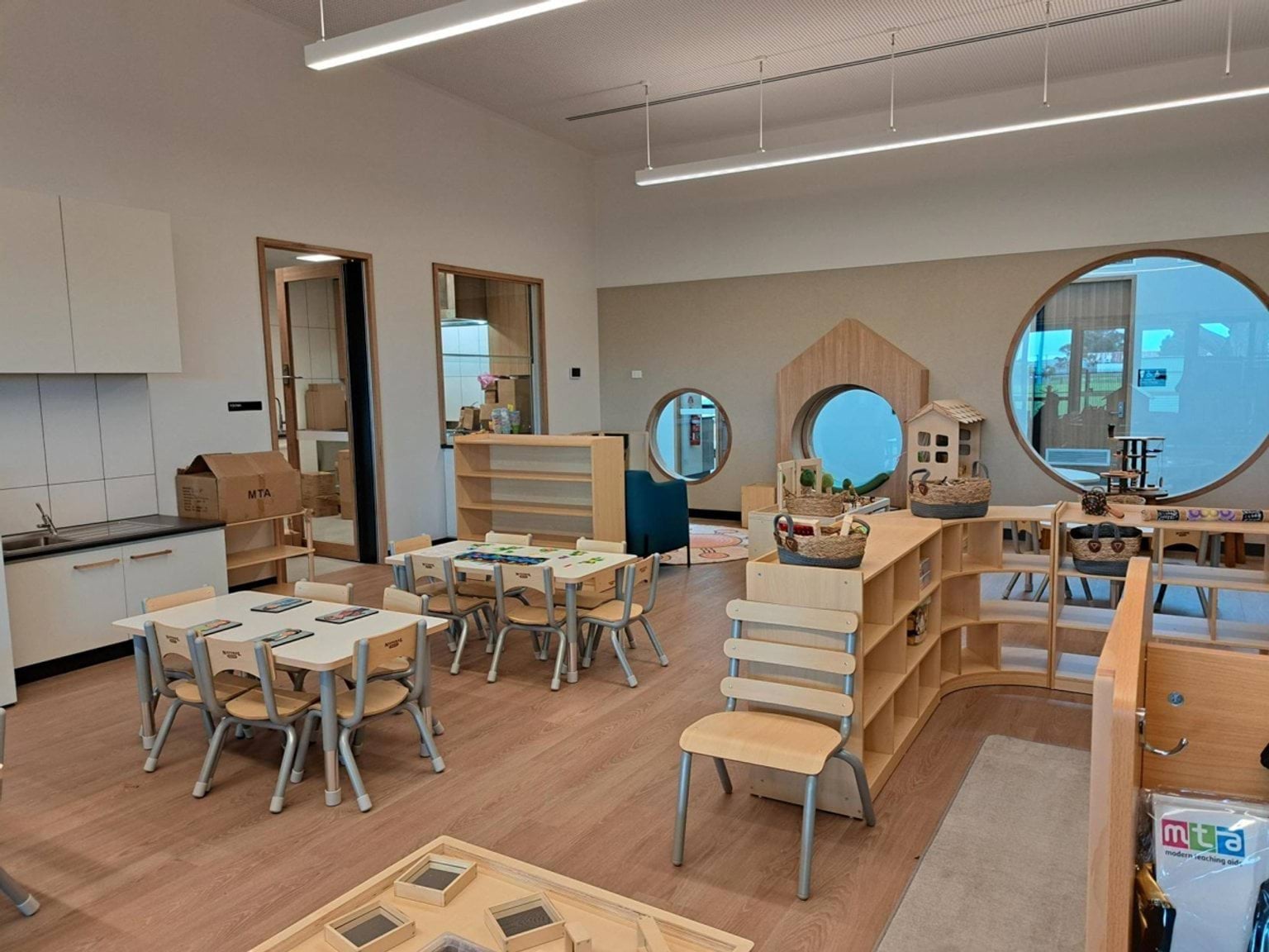 A kindergarten classroom with small tables, timber shelving, circular feature windows, and play equipment arranged around the room.