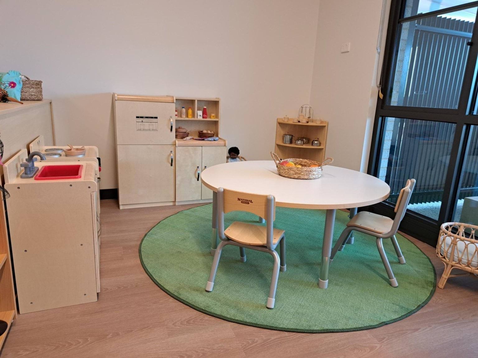 A dramatic play corner with a round green rug, a small table and chairs, and timber play kitchen units and storage shelves.