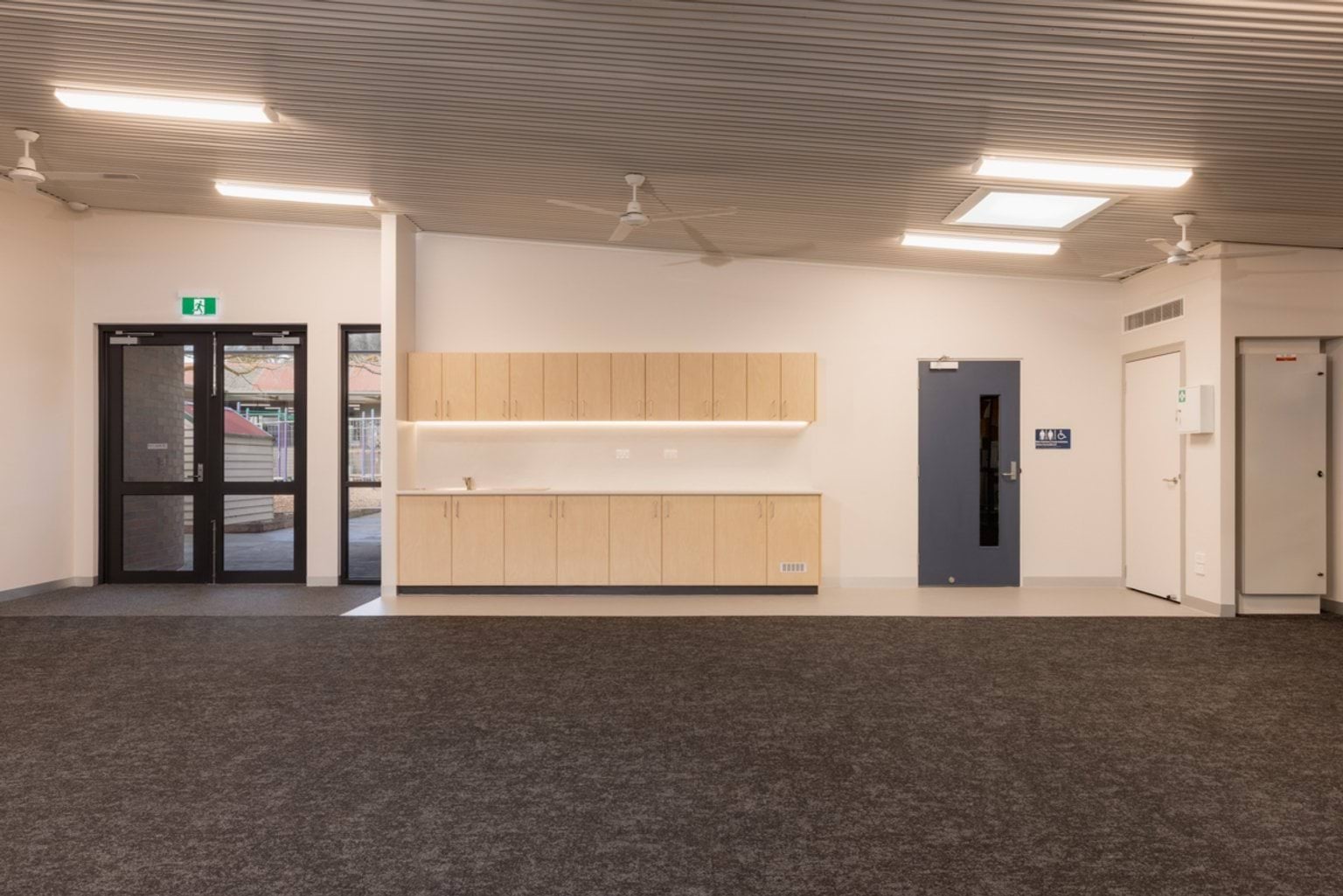 A spacious interior learning area with dark carpet, timber cabinetry, ceiling fans, and double glass doors leading outside.