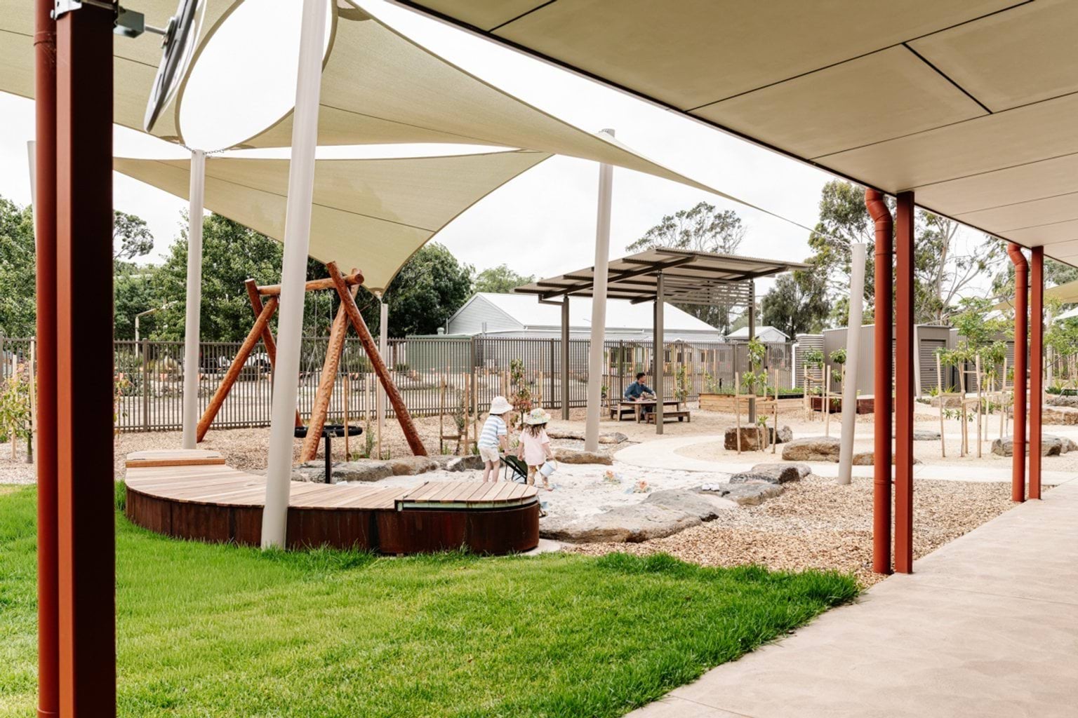 An outdoor play area with shade sails, timber decks, natural play elements, and green lawn viewed from a covered walkway.