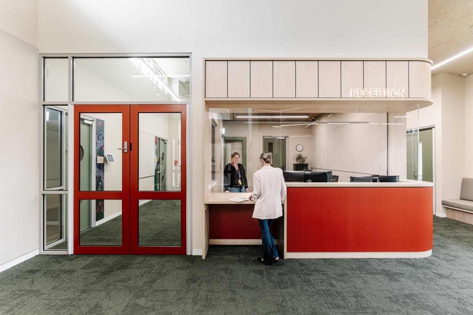 A reception area with a red and timber counter, glass screening, and a person speaking with staff behind the desk.