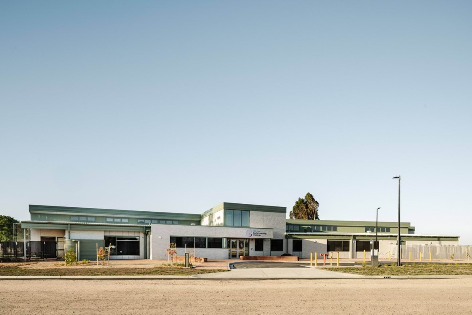 A wide exterior view of the school building with large windows, landscaped beds, and an open forecourt.