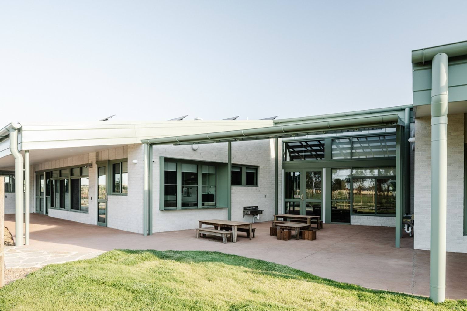 A covered outdoor learning area with timber tables and benches beside large windows into the school building.