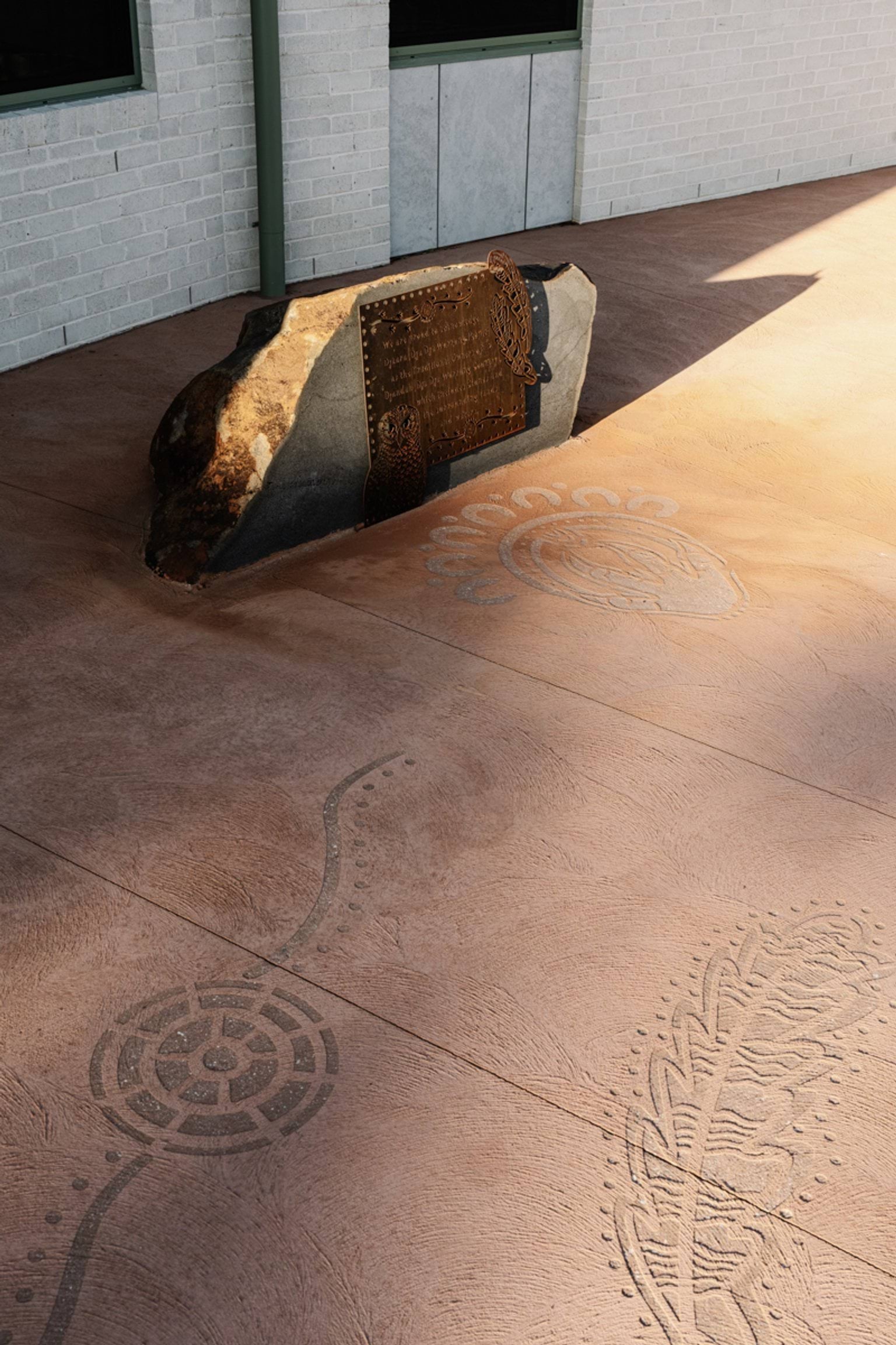 Etched patterns in the concrete near a large boulder with an engraved metal plaque outside the school building.