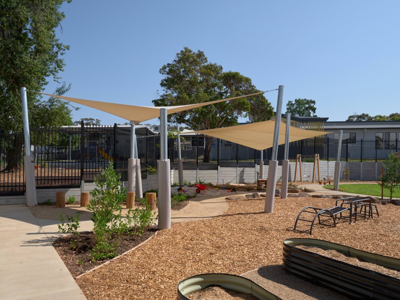 An outdoor play area with shade sails, natural landscaping, timber stepping logs, and play equipment on soft‑fall mulch.