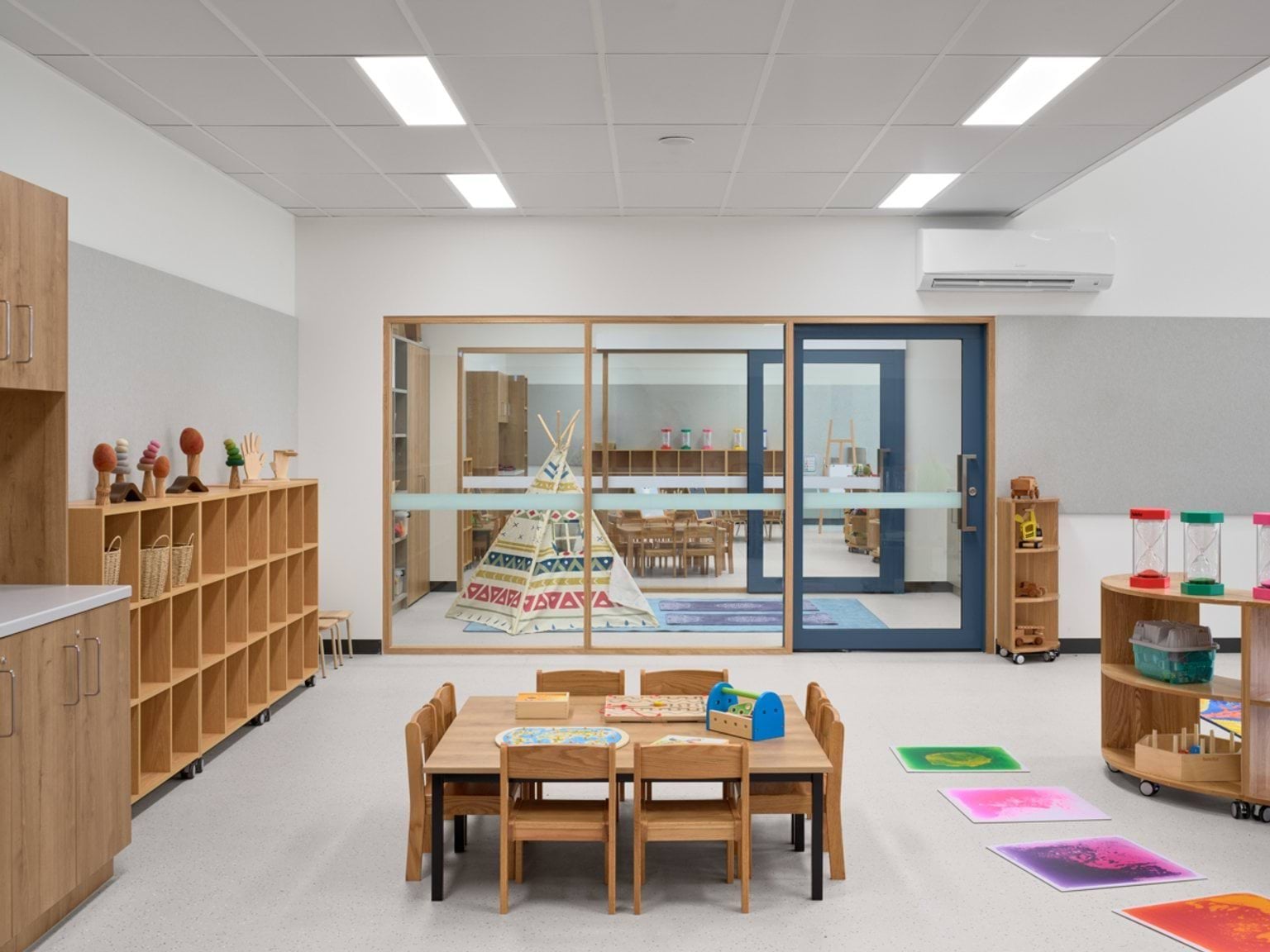 An early learning classroom with wooden tables, shelving, a play teepee, and a large internal window looking into an adjoining learning space.