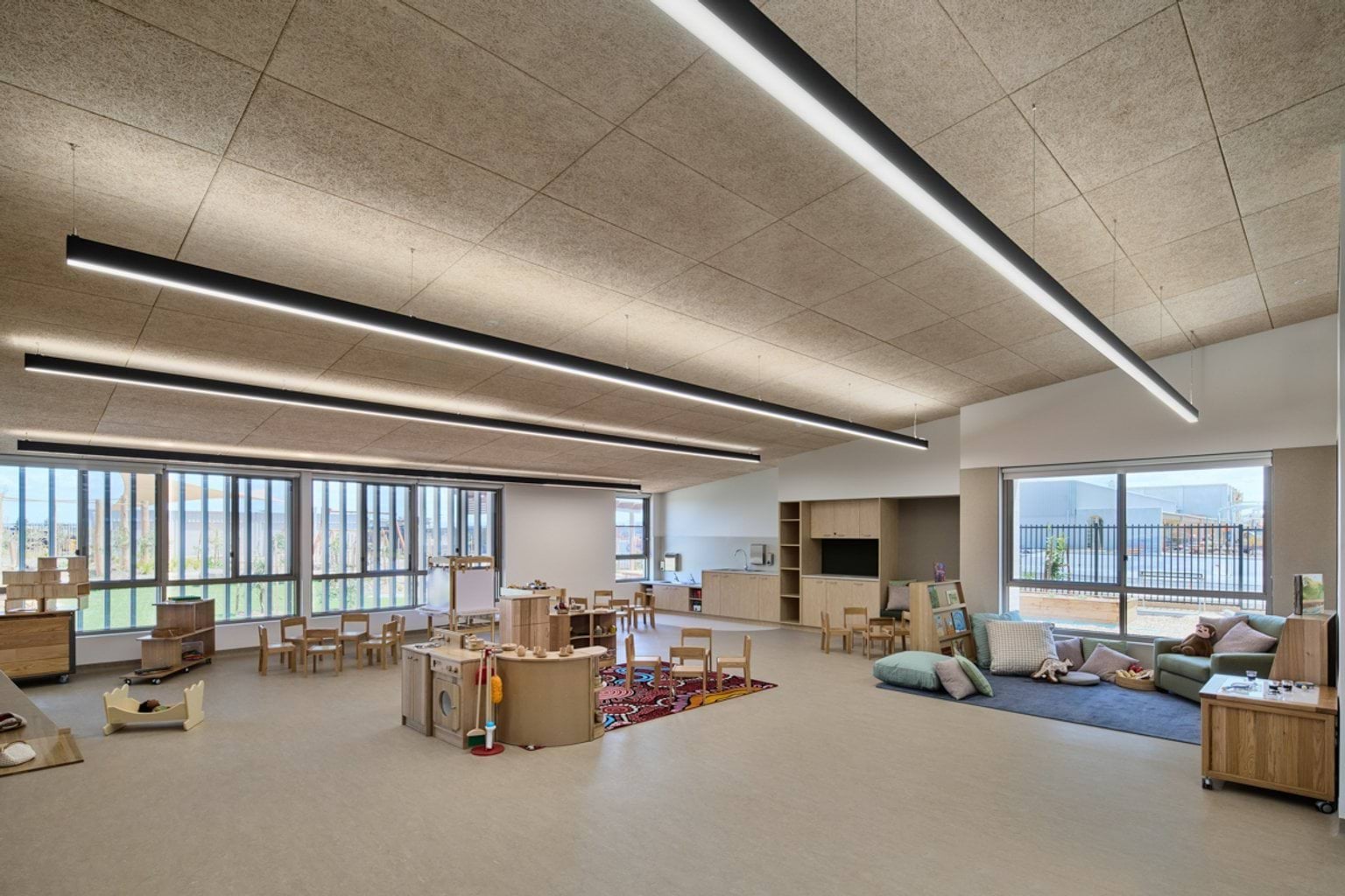 A spacious learning room with large windows, play stations, furniture, and a soft reading area with cushions.