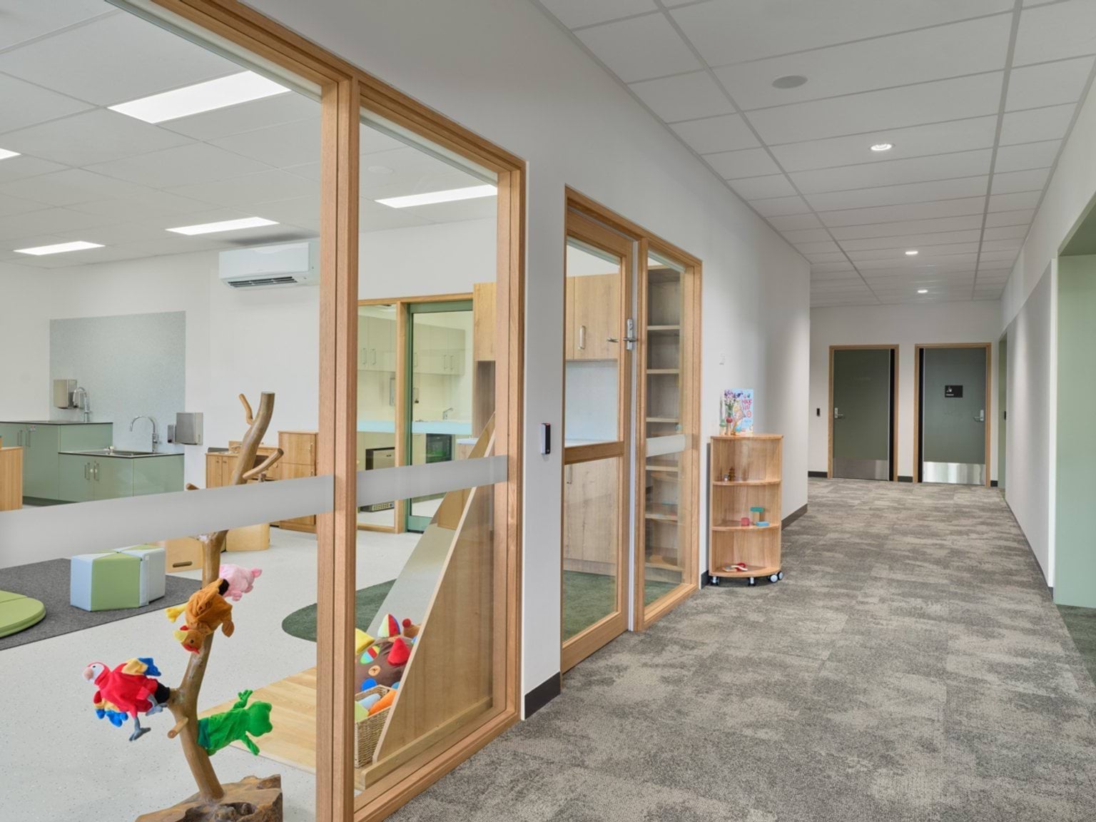 A hallway with carpeted flooring and glass windows overlooking indoor learning spaces, with shelves holding toys and books.