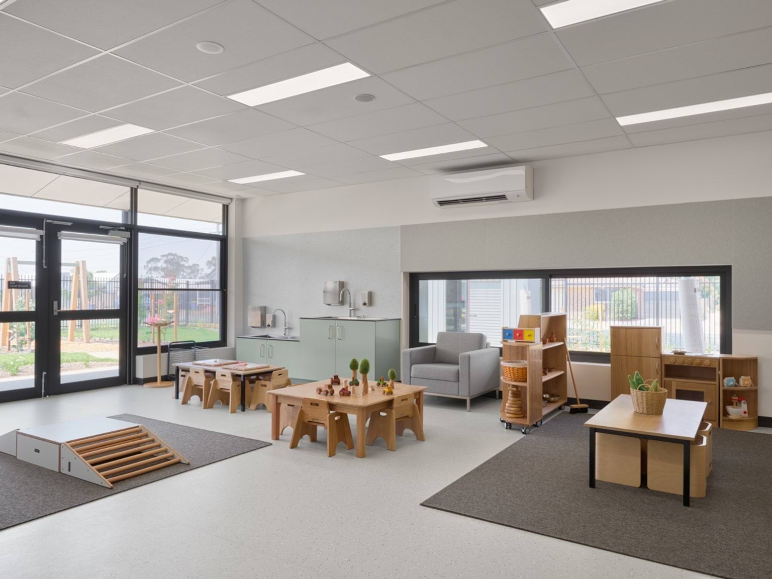 An early learning room with small tables, soft play mats, a reading corner, and views of the outdoor play area.