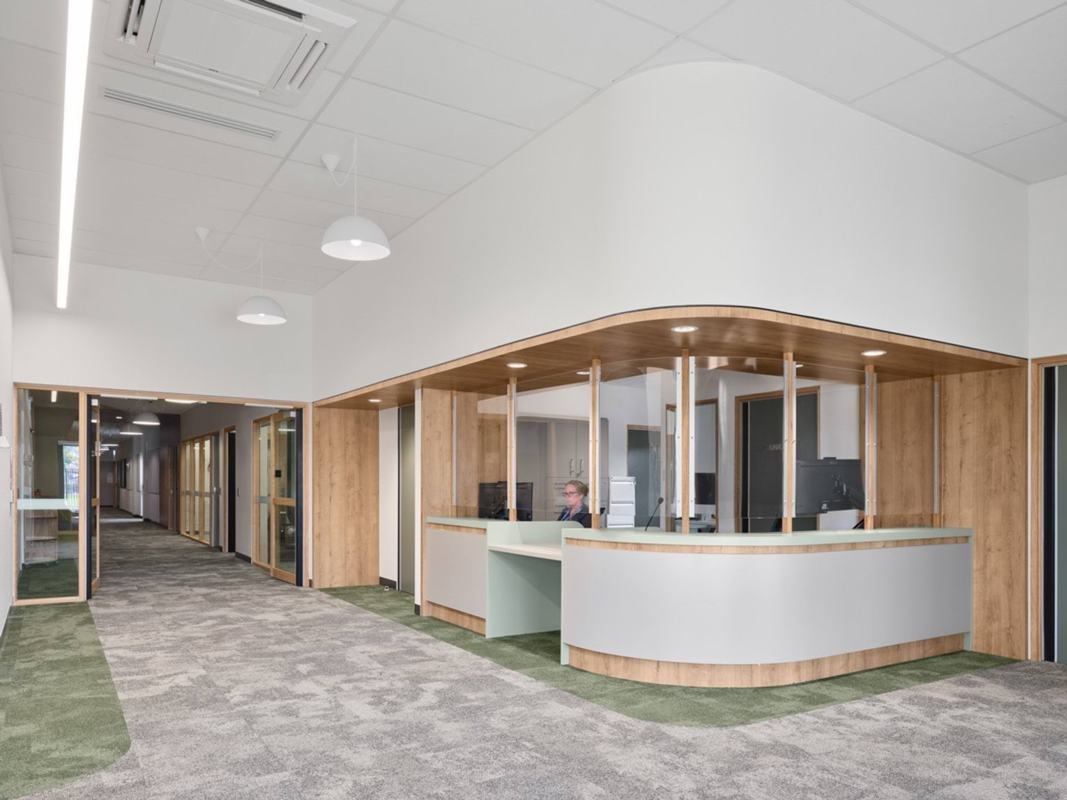 A reception area with a curved timber and white counter, carpeted hallway, and glass offices along the corridor.