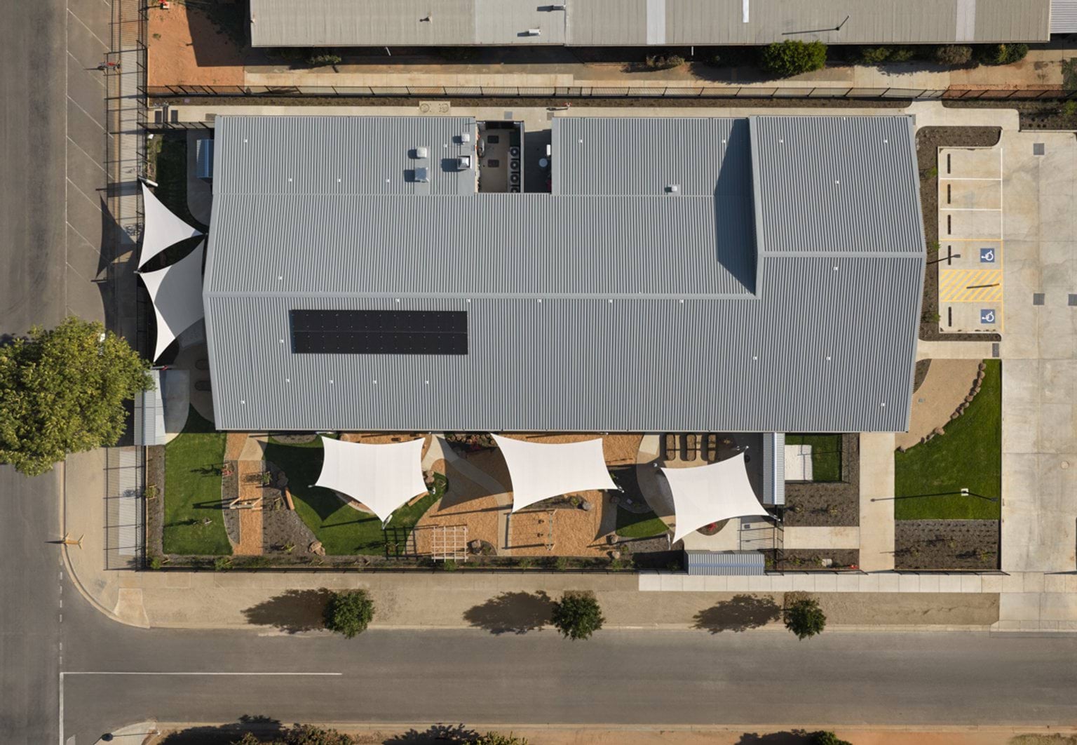 An overhead photo of the completed Early Learning Victoria Numurkah centre. There is a grey roof with some solar panels, white shade sails in the surrounding garden and playground, and a car park with 3 standard and 2 disabled parking spaces visible. 