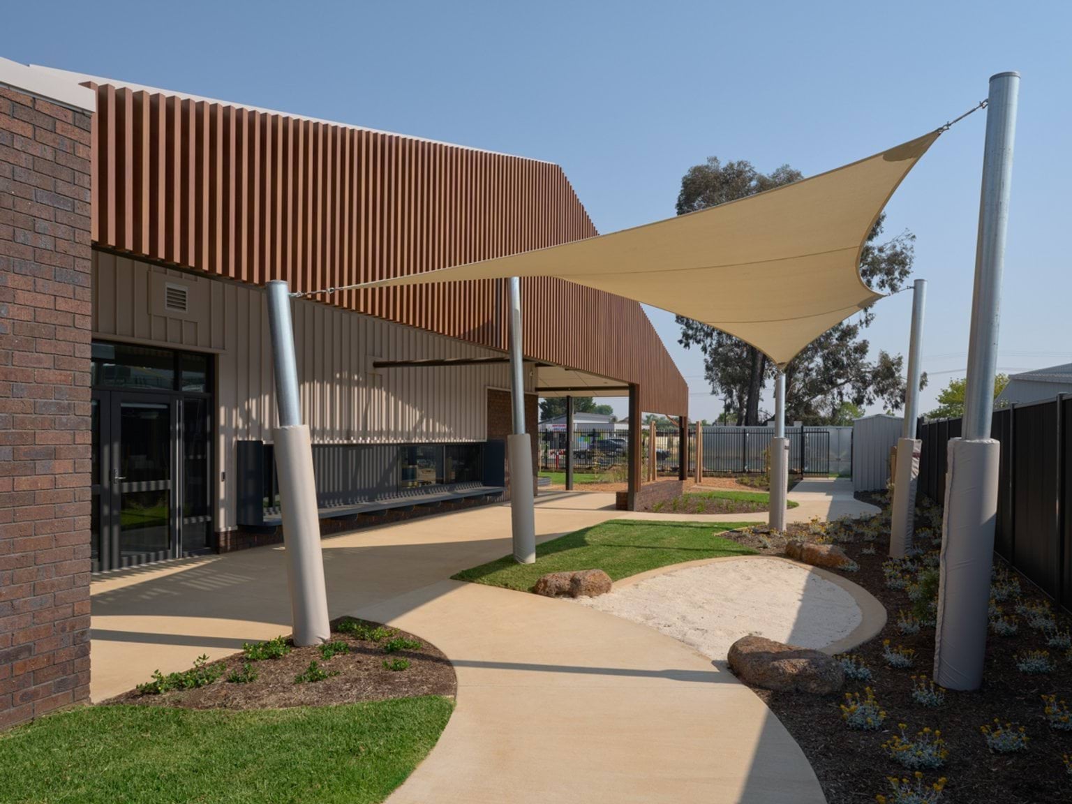 An exterior walkway at the early learning centre featuring curved paths, shade sails, garden beds, and timber‑screen cladding.