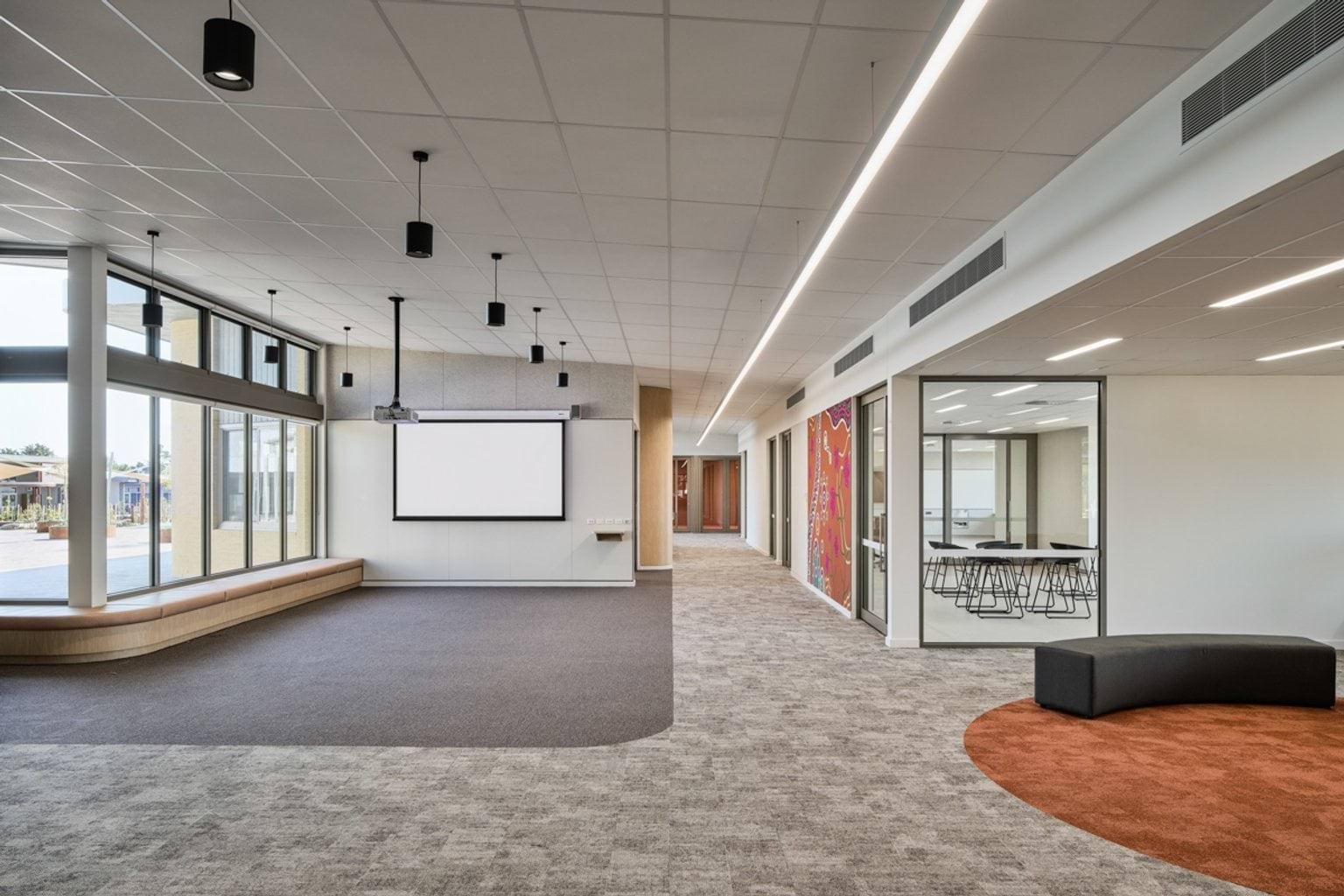 An open indoor learning area with a projector screen, large windows, and adjoining glass‑fronted meeting rooms.