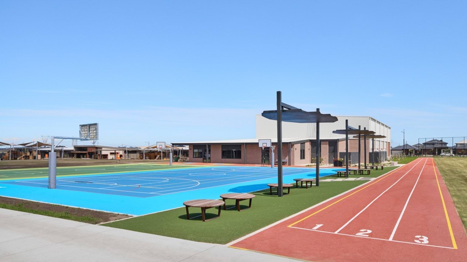An outdoor sports area with bright blue courts, shade structures, circular benches, and a red running track beside the building.