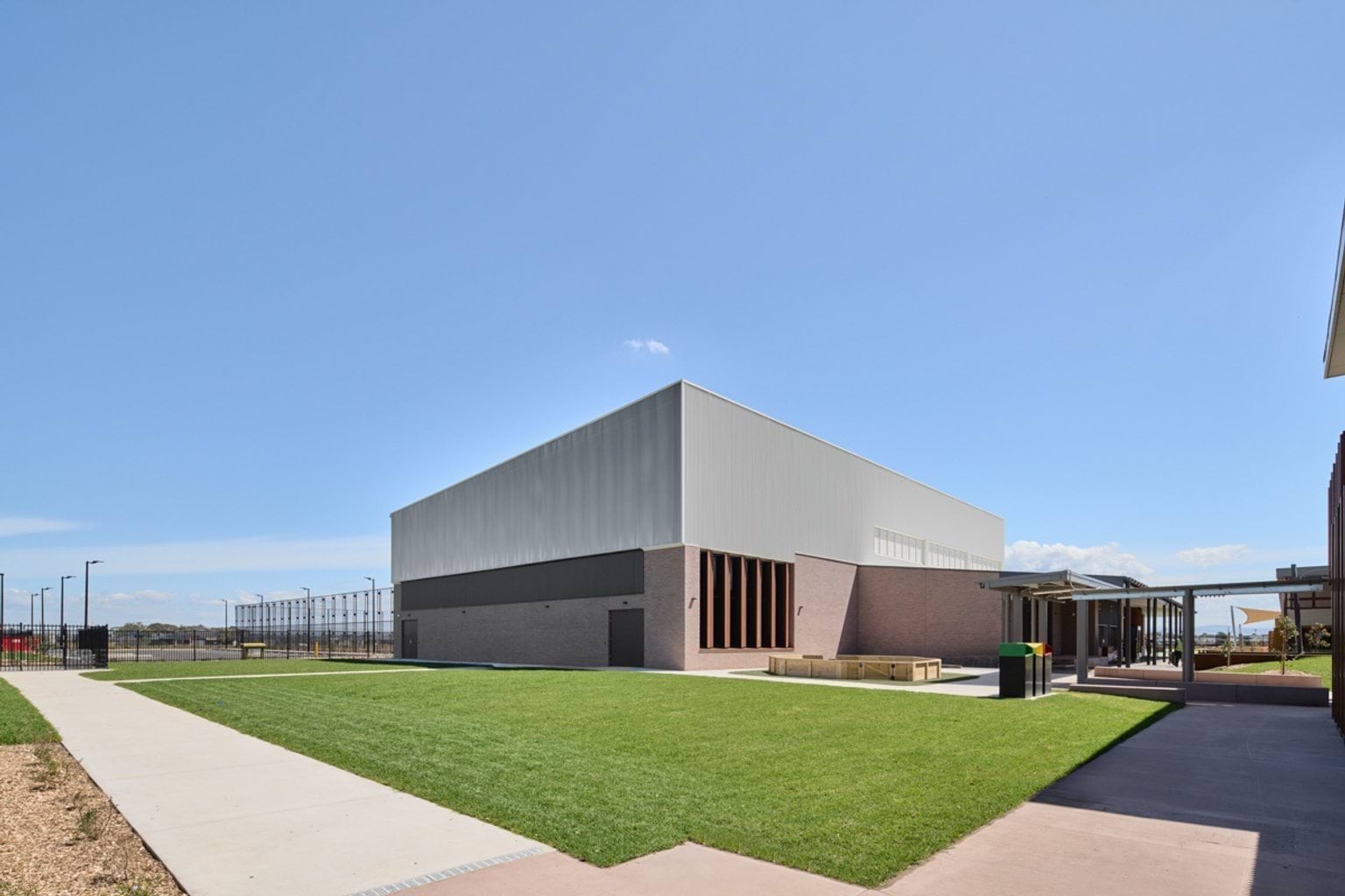 A large open lawn area beside a tall school sports building with brick and metal cladding.