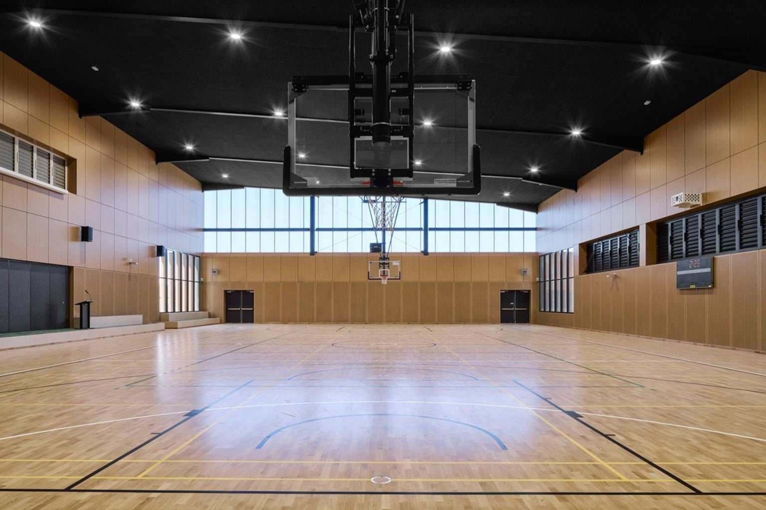 An indoor sports court with a timber floor, high ceiling, large windows, and a basketball hoop at the centre.
