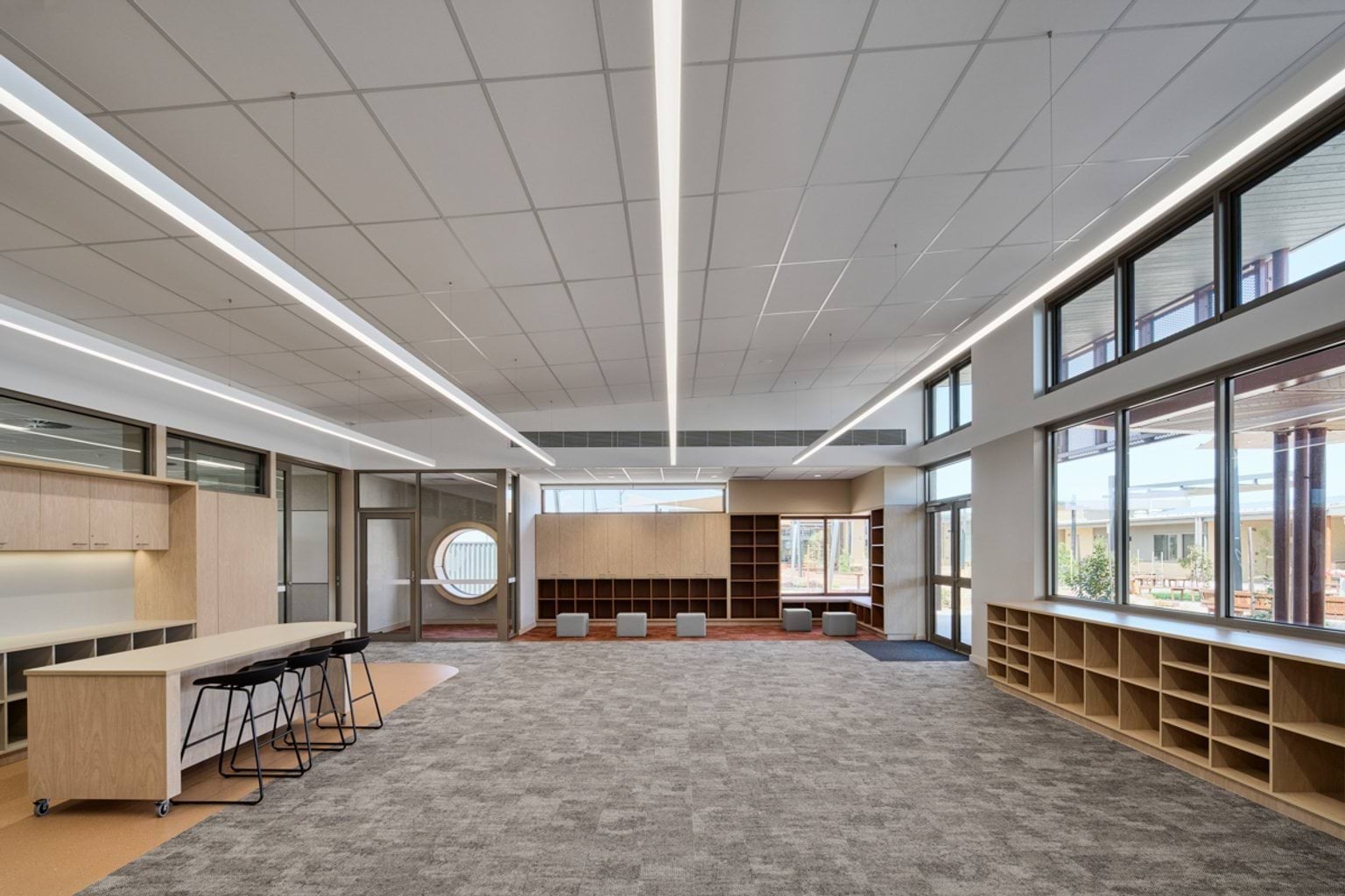 A spacious learning area with carpeted flooring, built‑in shelving, large windows, and a central workbench with stools.