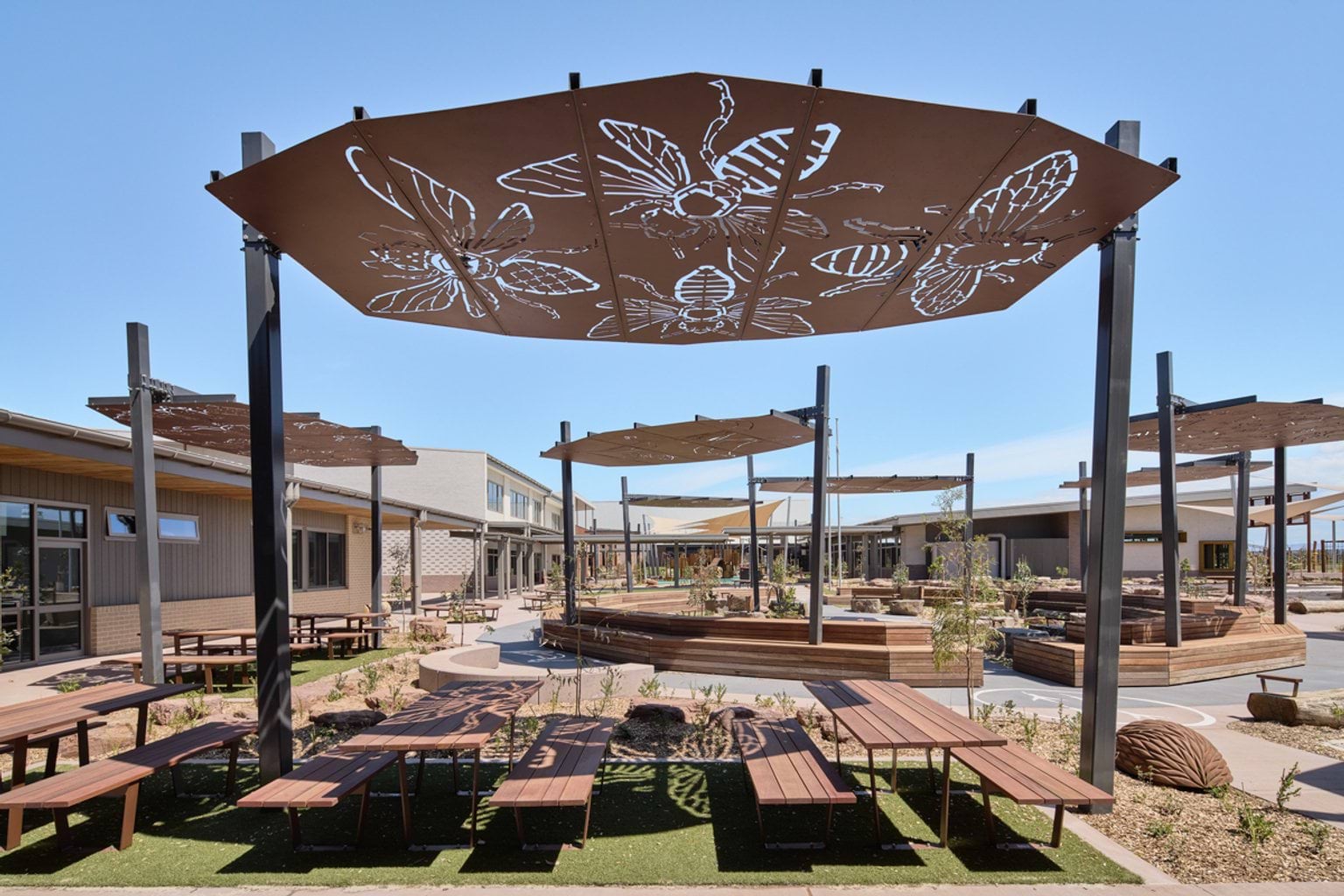 An outdoor seating and play area featuring metal shade structures with nature‑themed cut‑outs and timber benches.