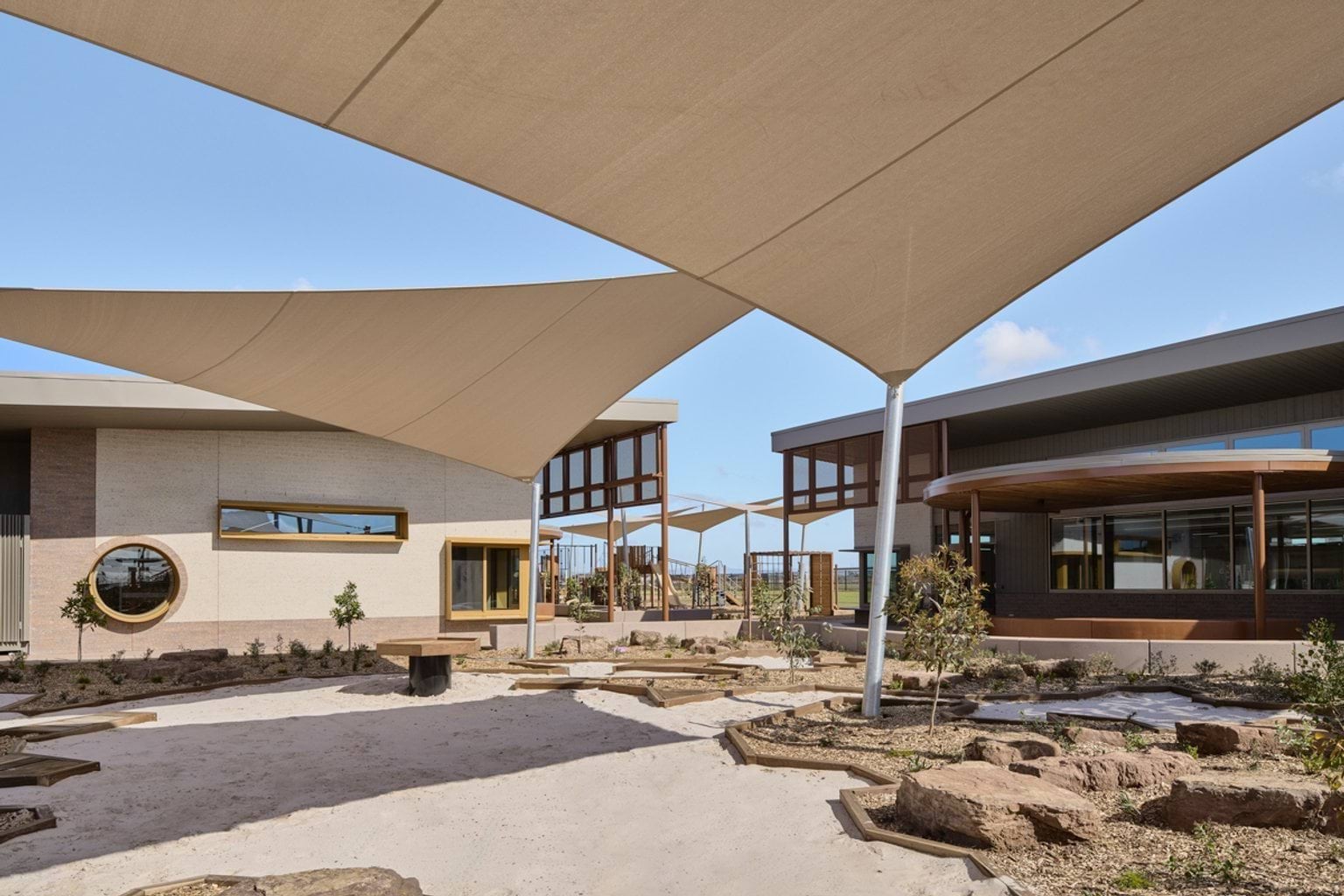 An outdoor play area with sand, shade sails, garden beds, and connecting paths between school buildings.