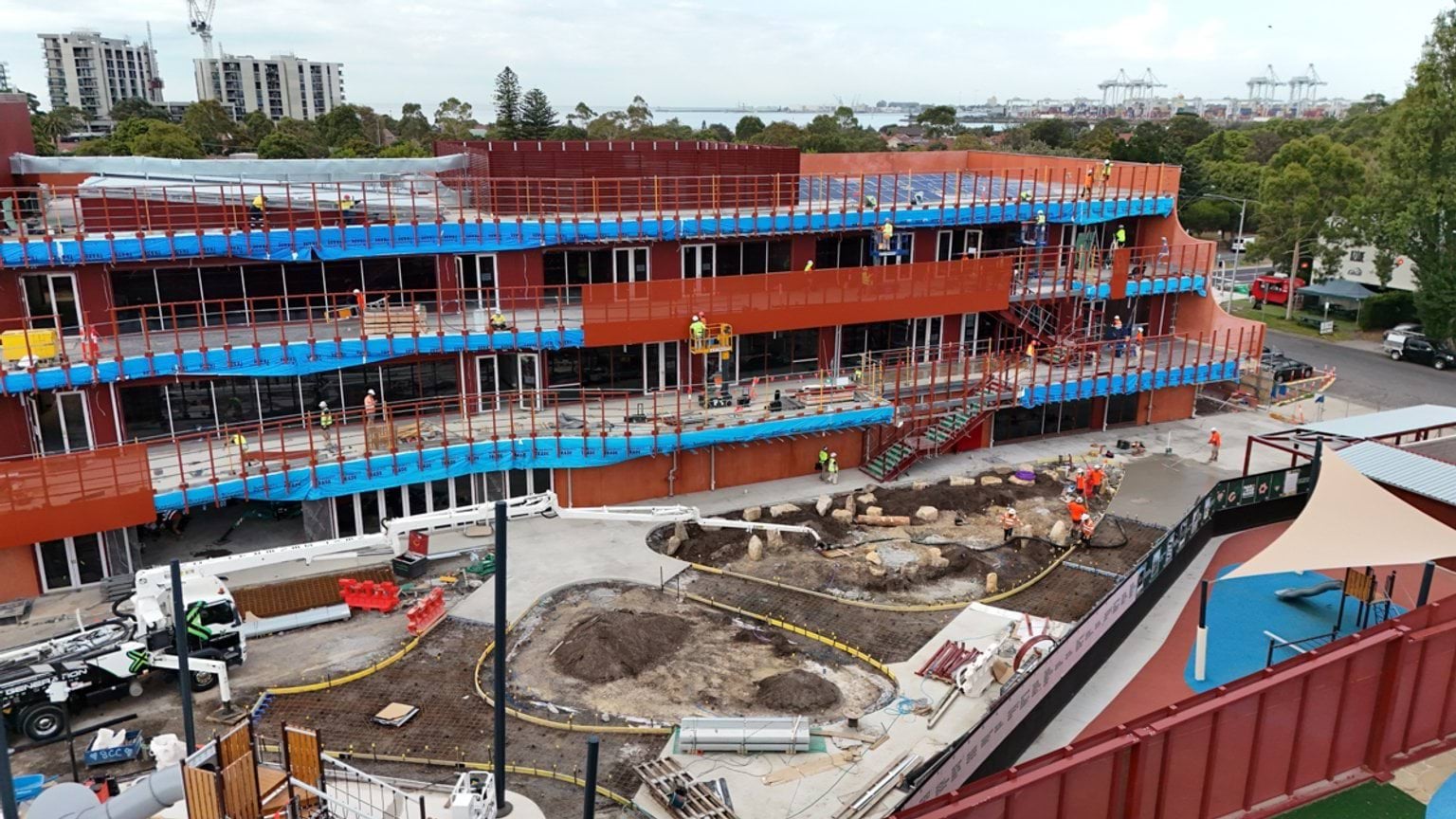 A construction site view of the school building showing red exterior panels, scaffolding, and landscaping works underway.