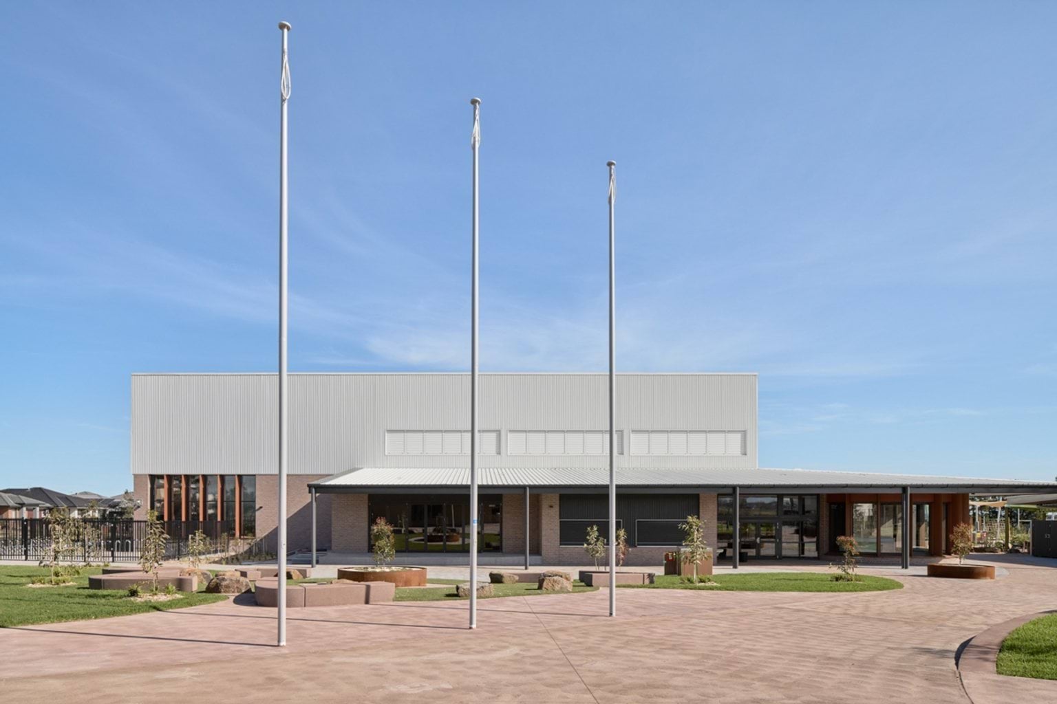 A ground‑level view of a school building with three flagpoles in front, a covered entry, and landscaped paths and garden beds.