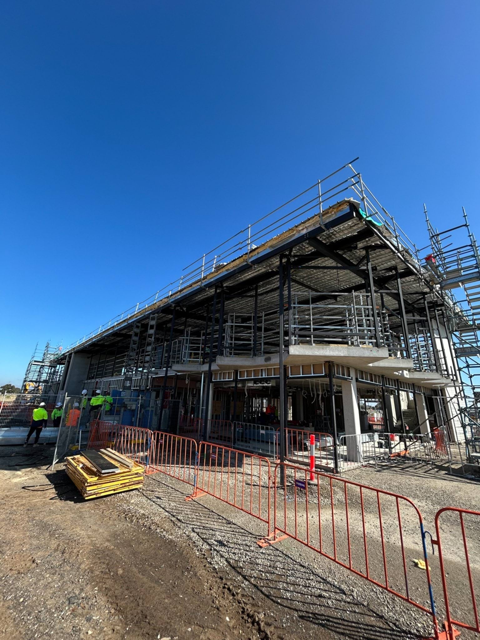 A partially constructed two‑storey school building with scaffolding, roof framing, and workers on site, surrounded by temporary fencing and construction materials.