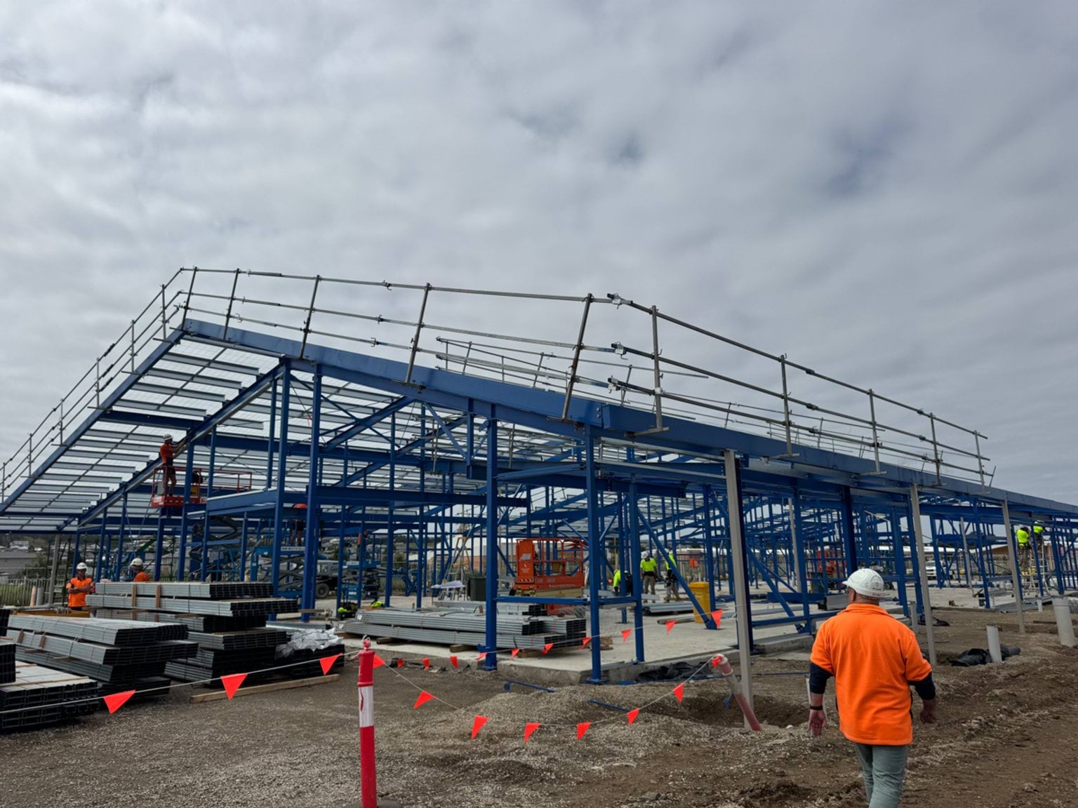 A steel-framed school building under construction, with workers on site and materials stacked around the structure.