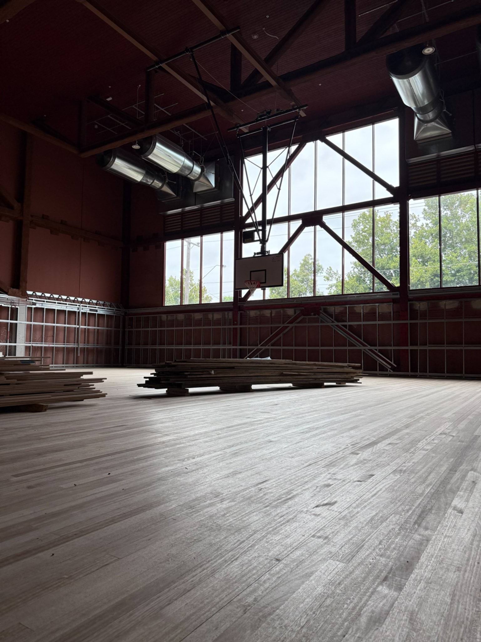 A large sports hall with timber flooring being installed, exposed roof trusses, and a basketball hoop mounted on the wall.
