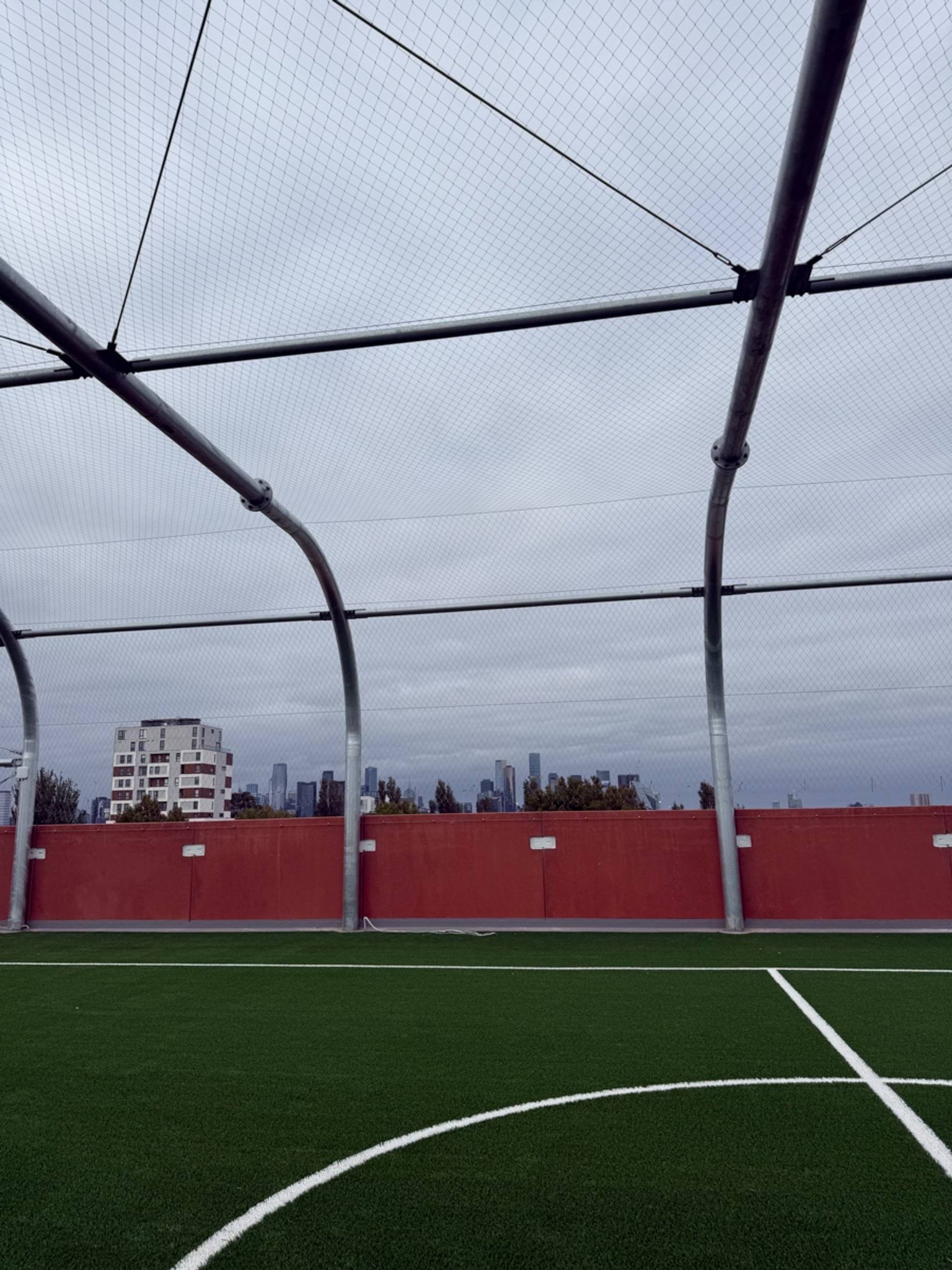 A rooftop sports court with synthetic turf and protective netting overhead, with city buildings visible in the distance.