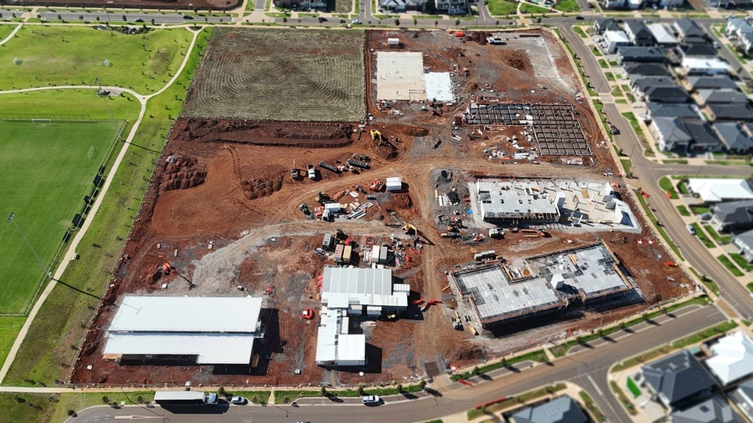 Elevated aerial view showing several school buildings under construction, with concrete slabs, construction vehicles, and surrounding residential streets.