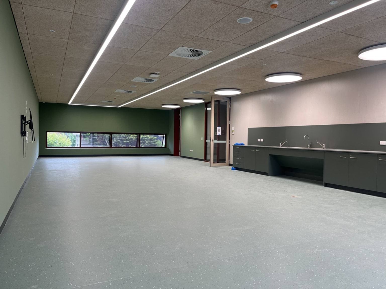 A large multi‑purpose learning space with green feature walls, vinyl flooring, ceiling lights, and a kitchenette along one wall.