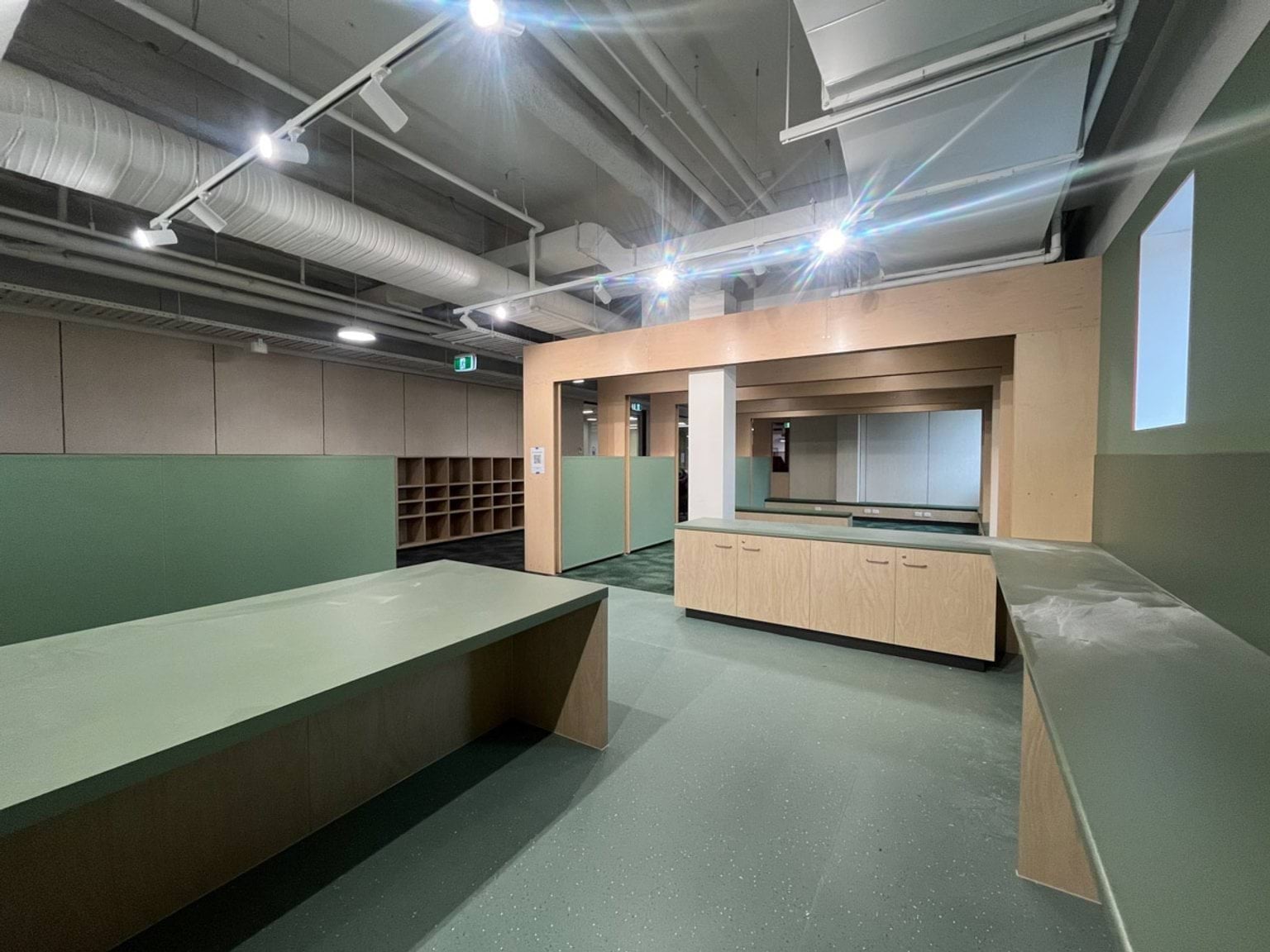 A practical learning space with timber benches, storage cupboards, green work surfaces, and exposed ceiling ductwork.