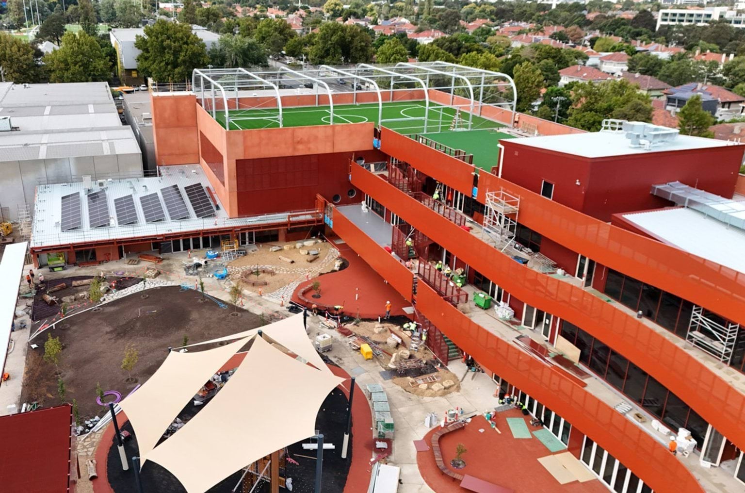 Elevated view of the school showing rooftop sports courts with synthetic turf, surrounding buildings, and construction activity below.