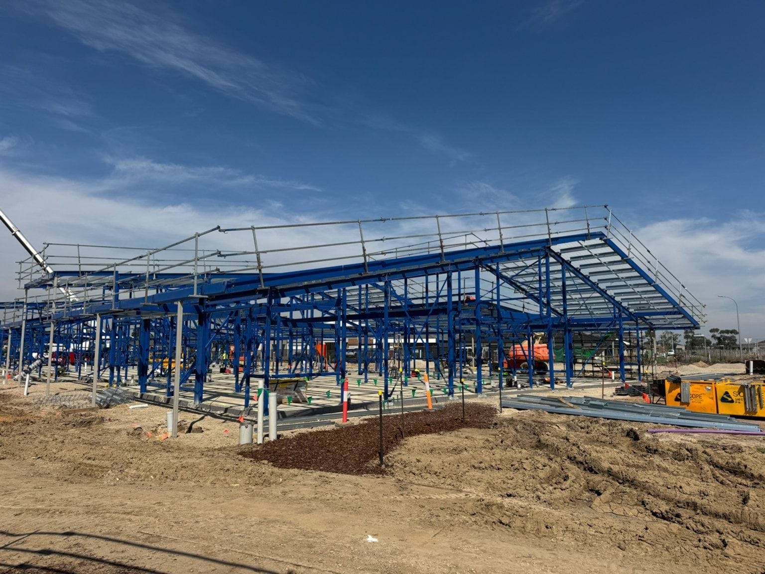 A large steel structure forming part of a school building, with roof framing in place and foundation works visible below.