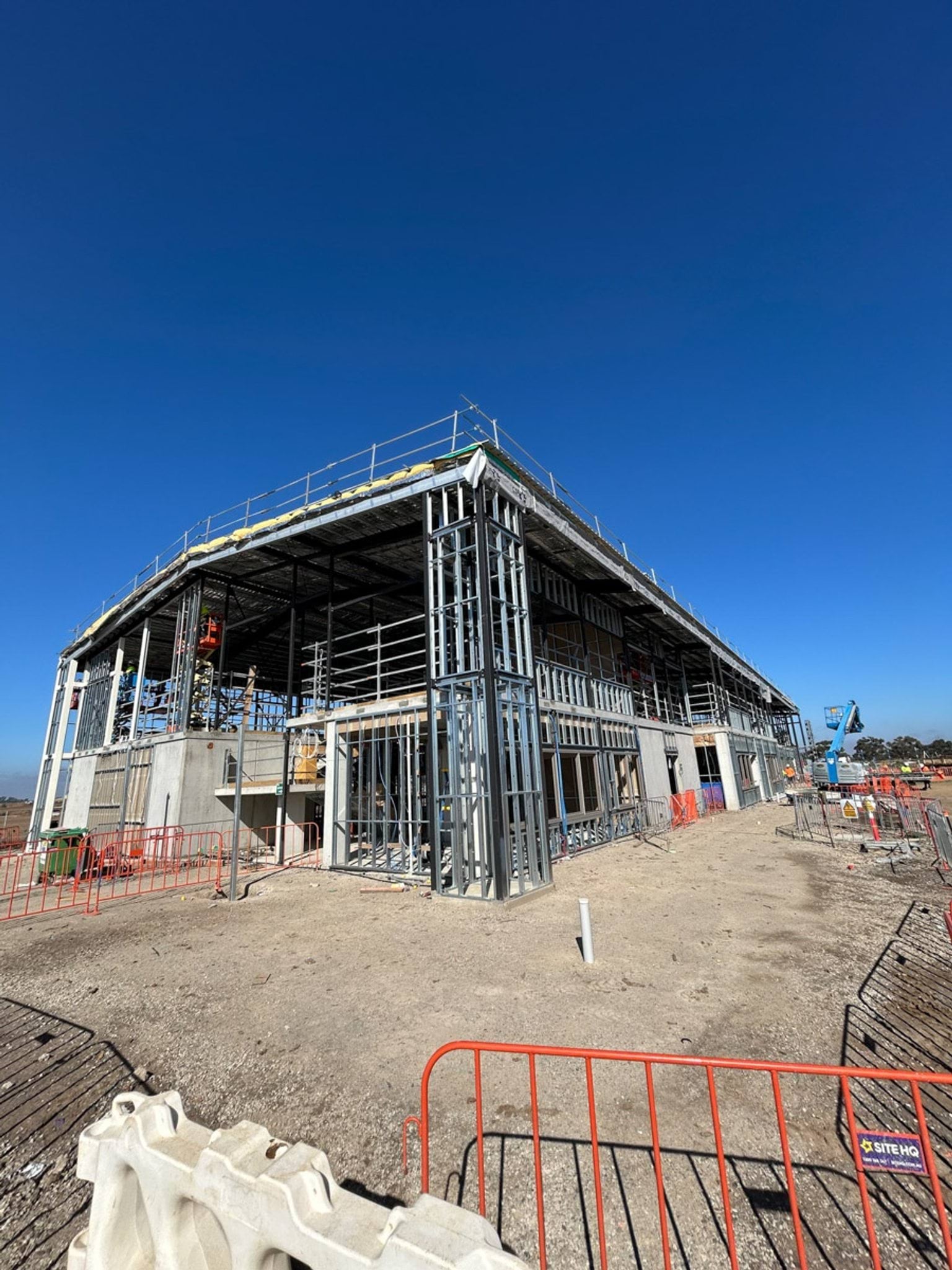 A two‑storey school building under construction with steel framing, scaffolding, and safety fencing on a cleared site.