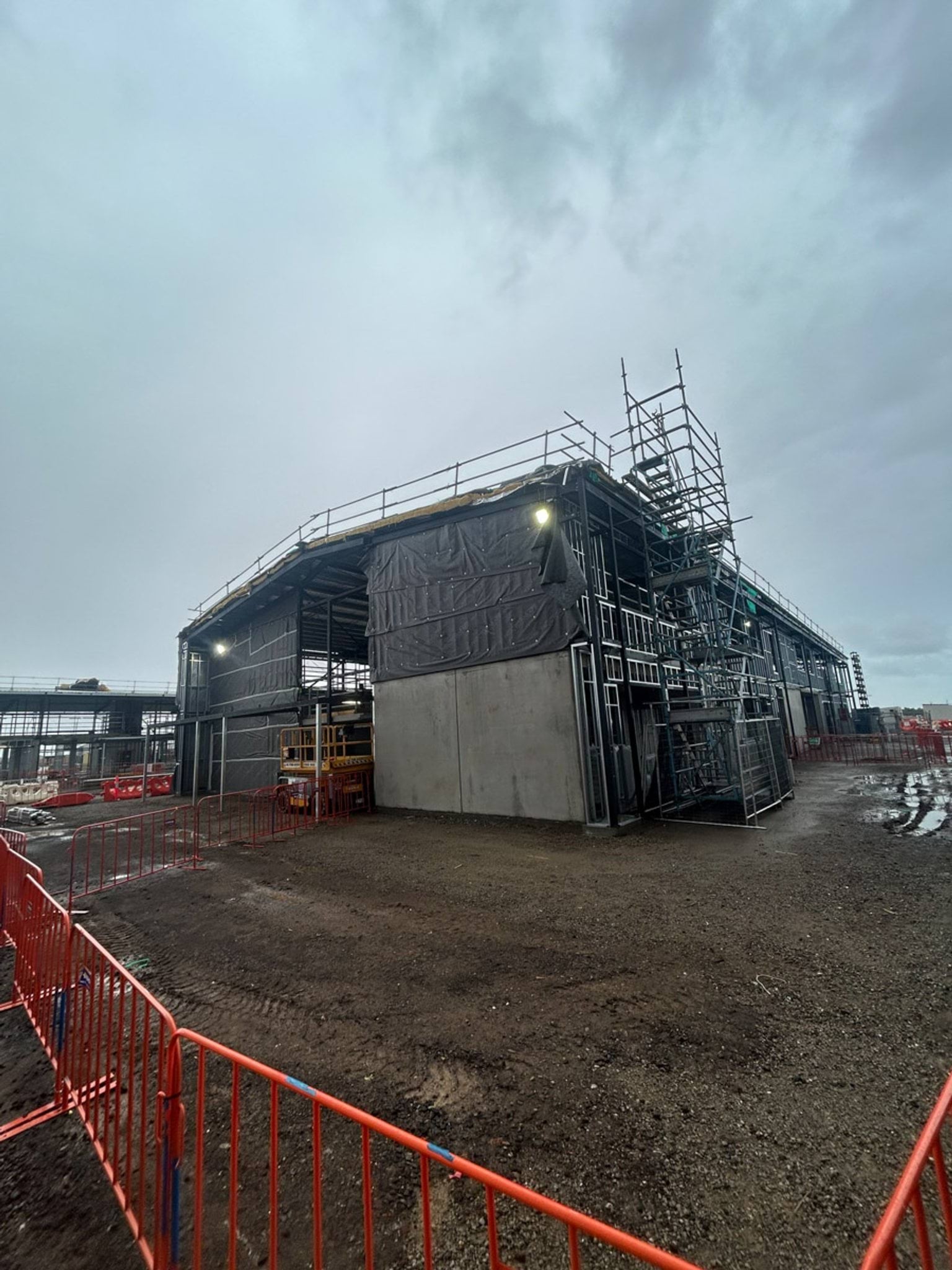 Corner view of a school building under construction with scaffolding, wrapped wall sections, and site lighting during overcast conditions.