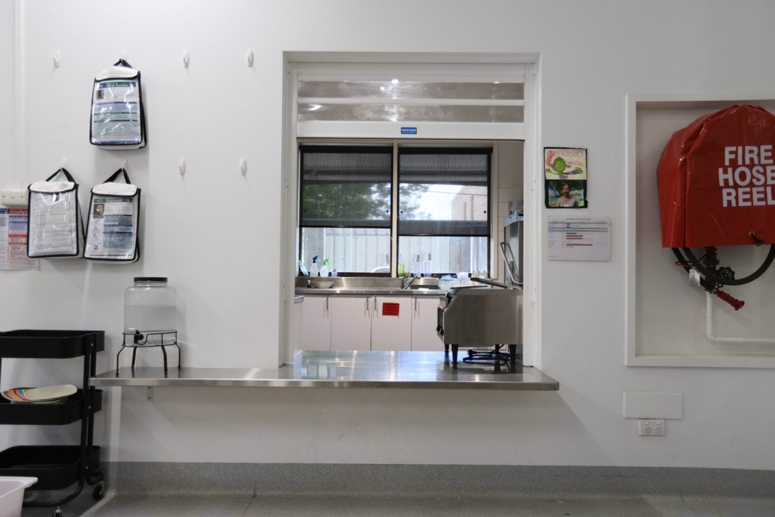 School kitchen servery window with stainless steel counter, water dispenser, and view into food prep area.
