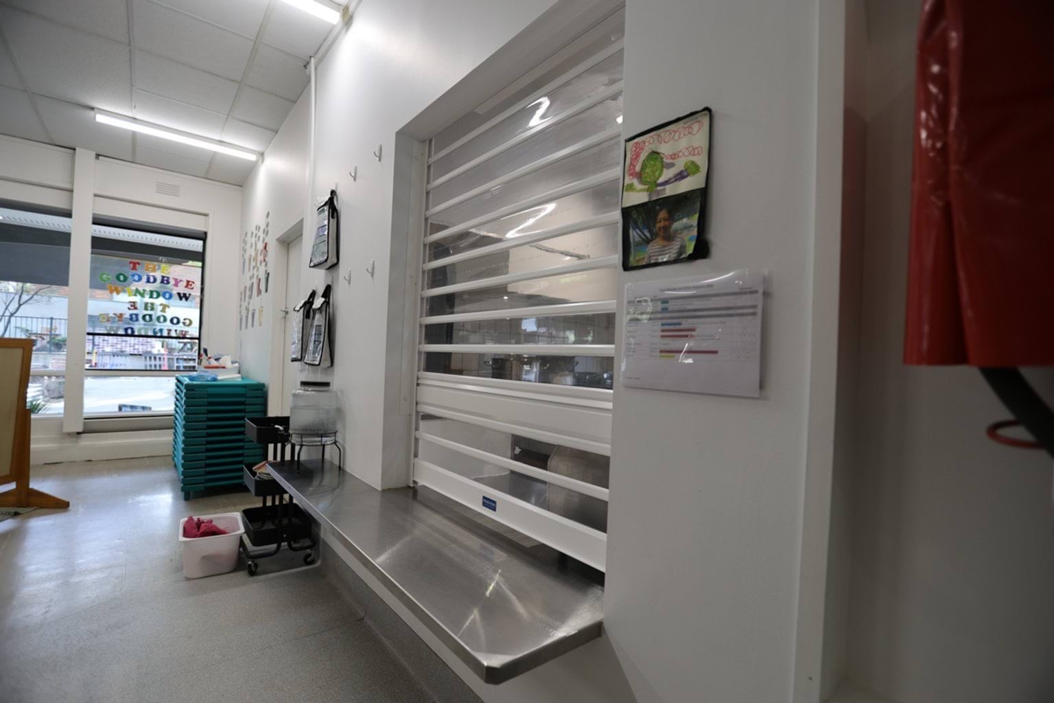 Closed servery shutter in a school kitchen, with stacked trays, trolley, and window to an adjacent learning space.