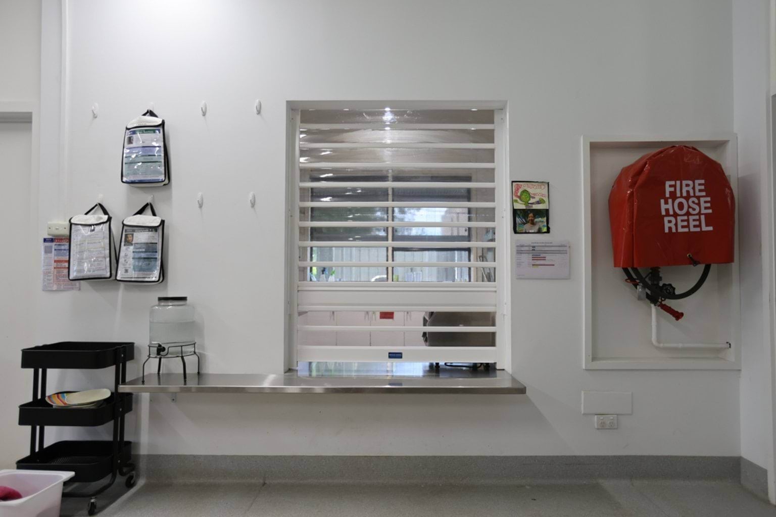 School kitchen servery with shutter partially down, stainless steel bench, wall-mounted documents, and fire hose reel nearby.