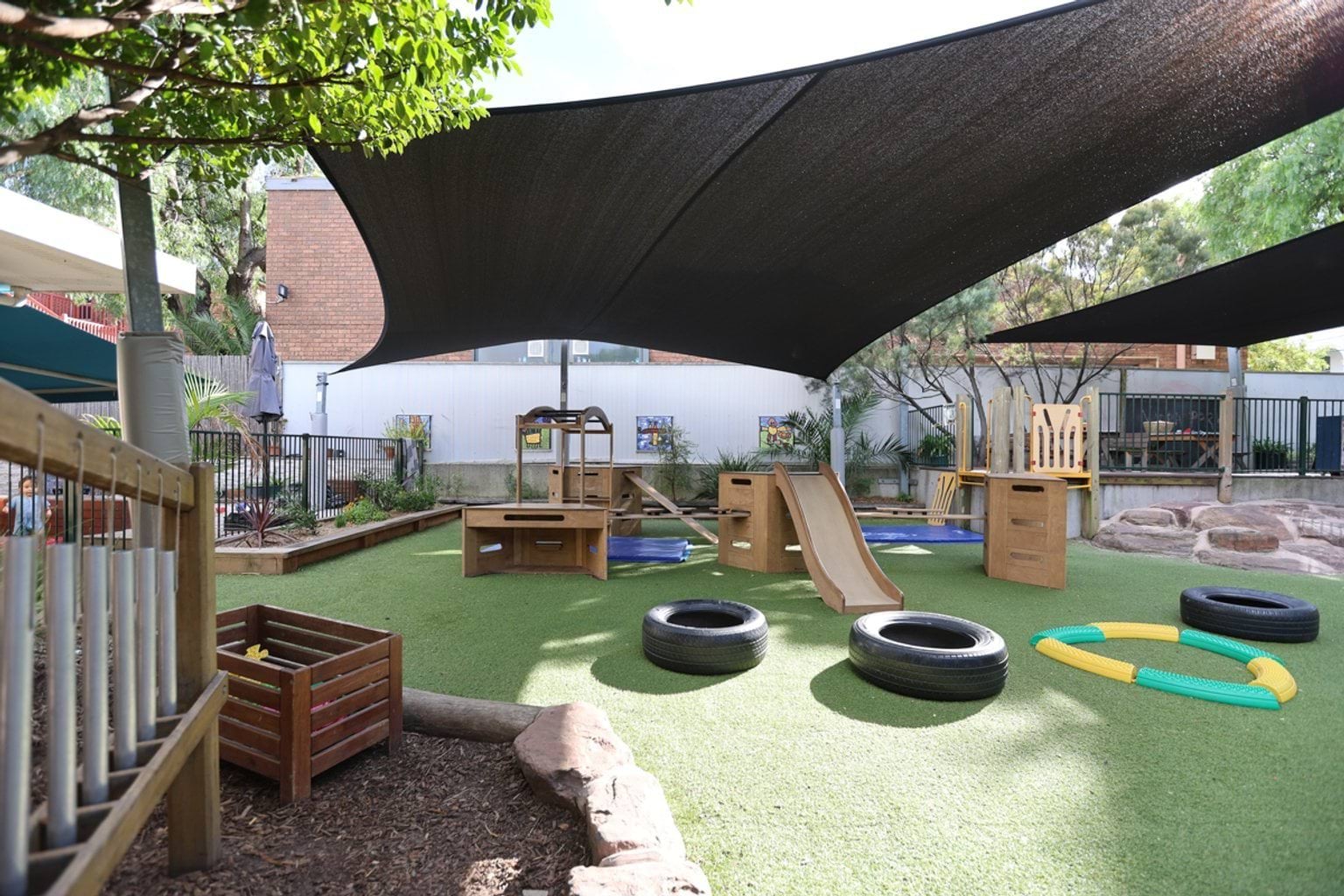 Outdoor play area showing slide, play tyres on ground, shade cloths, and school buildings in background.