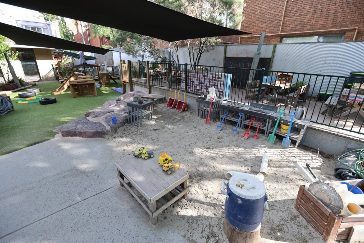 Sand play zone with digging tools, water trays and toy trucks beside a rock edge and shaded playground.