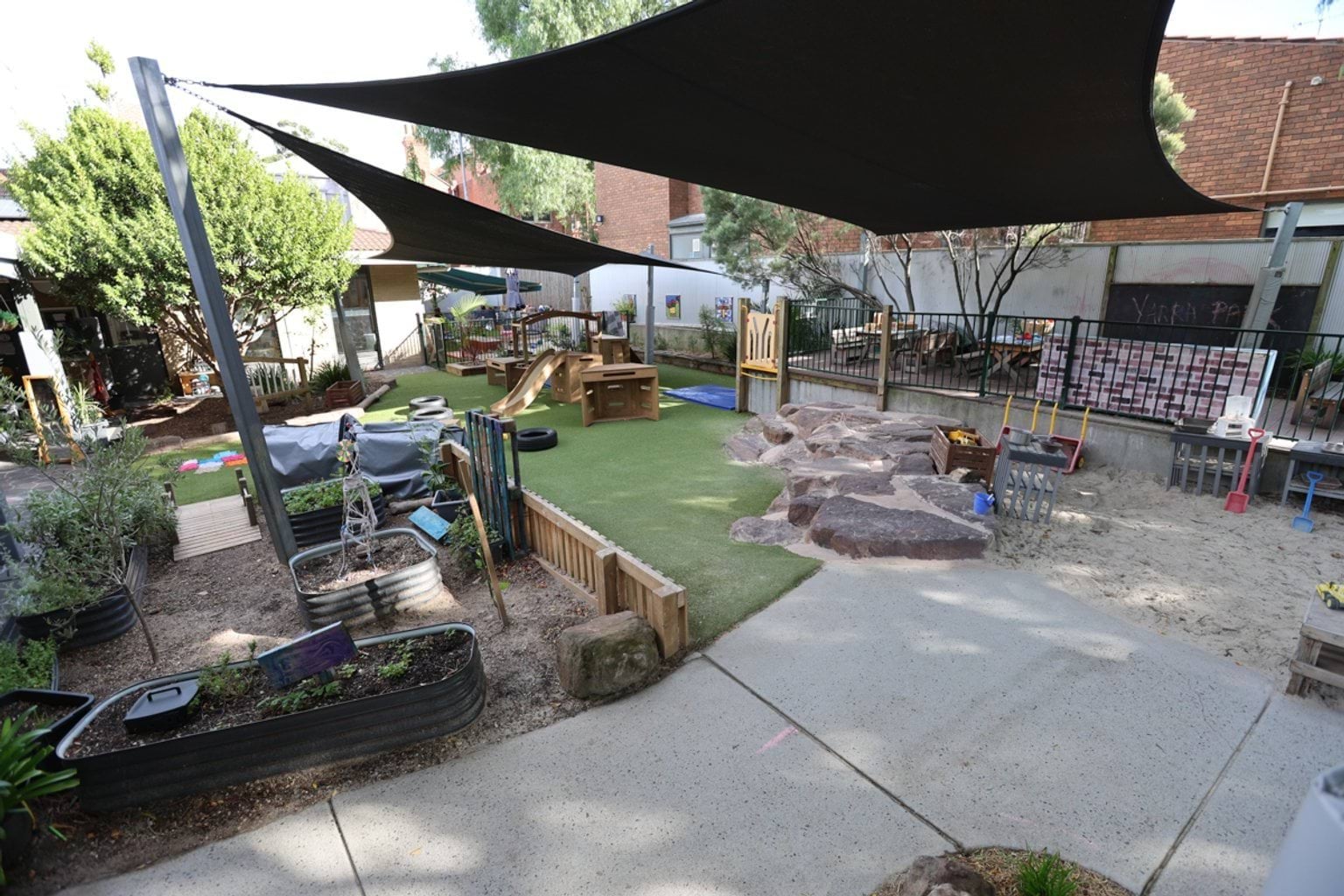 Shaded school playground combining garden beds, rock features, sand play and timber climbing equipment.